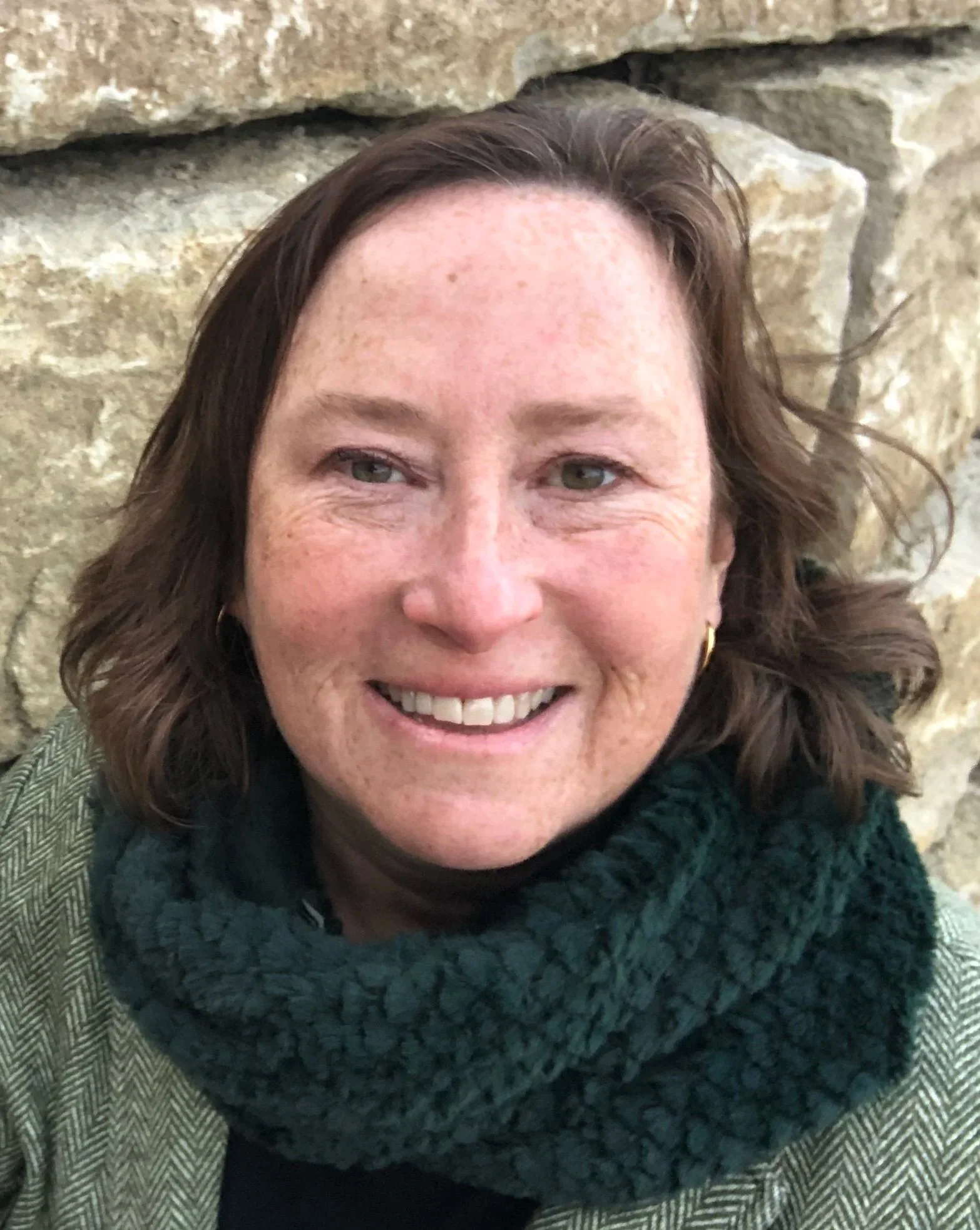 Close-up of Rachel, smiling, wearing a dark green scarf, and a patterned jacket, standing in front of a stone wall.