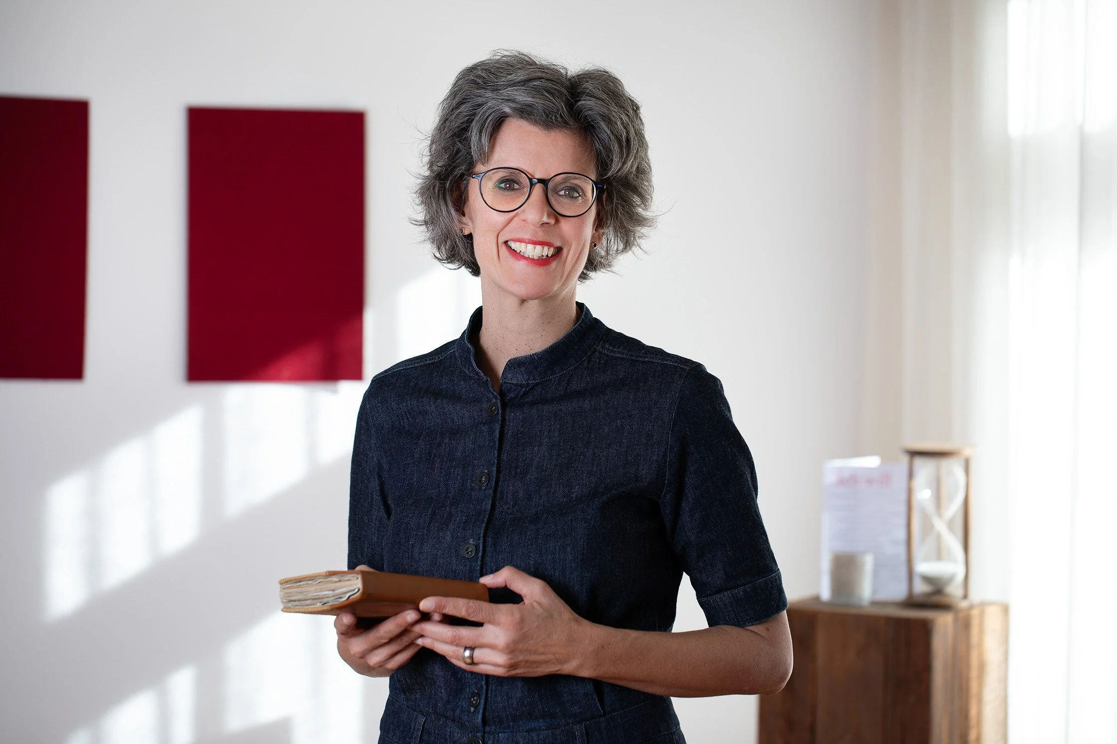 Lächelnde Frau mit Brille, kurzes graues Haar, dunkles Hemd, hält ein Notizbuch.