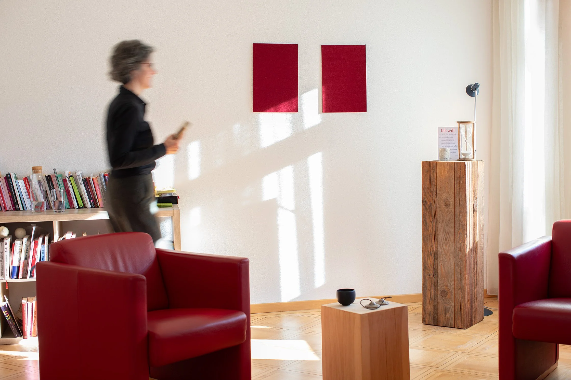 Ein Wohnzimmer mit rotem Sessel, Holzboden, Bücherregal, Standuhr, Wanddeko, und einer Person, die an einem Smartphone steht.