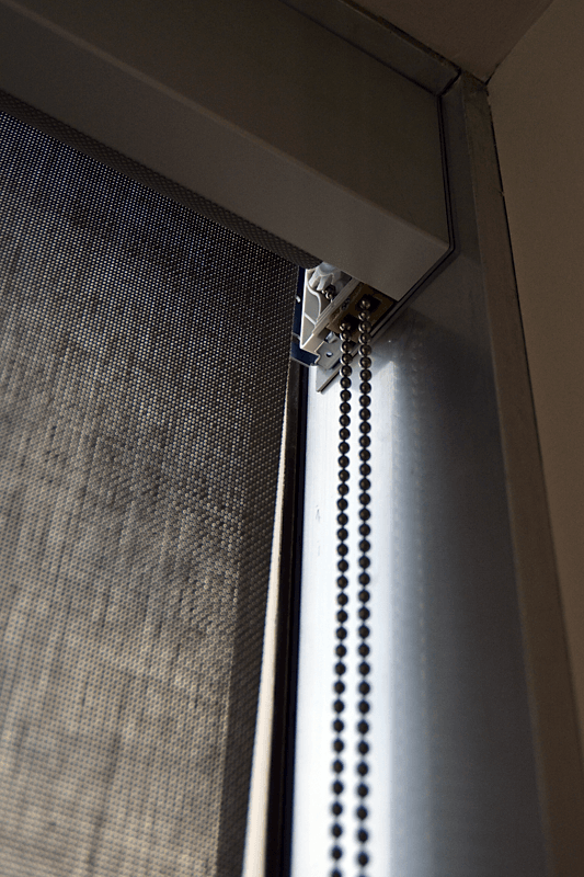 Close-up of a commercial solar roller shade headrail and bead chain mechanism, showing the textured fabric and smooth manual operation system.