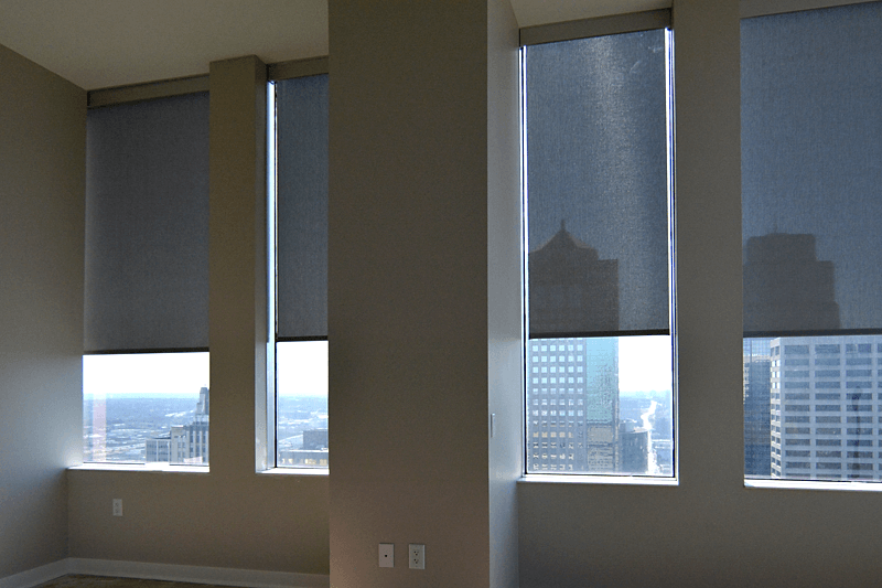 Three commercial solar roller shades partially lowered on tall office windows, filtering sunlight while preserving a city skyline view and reducing glare.
