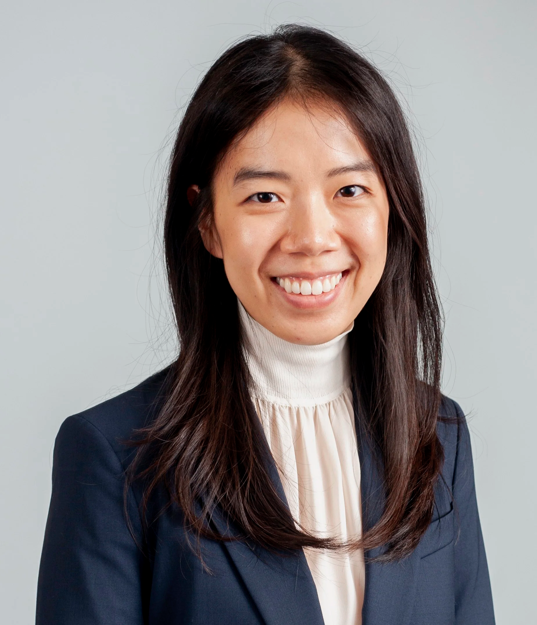 Headshot of a woman with long dark hair, smiling, wearing a dark blazer and a white turtleneck, against a plain light background.