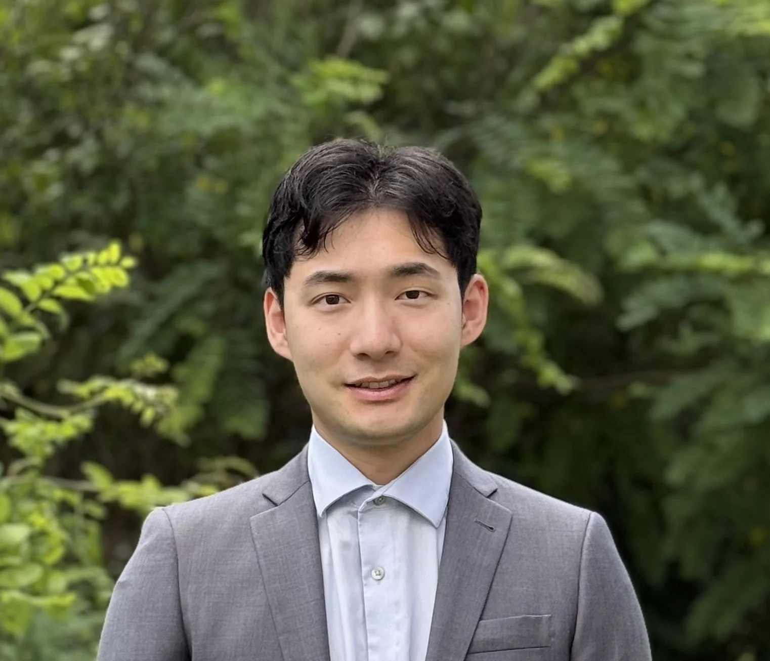 A young man with black hair wearing a grey suit and light blue shirt smiling outdoors with green foliage in the background.