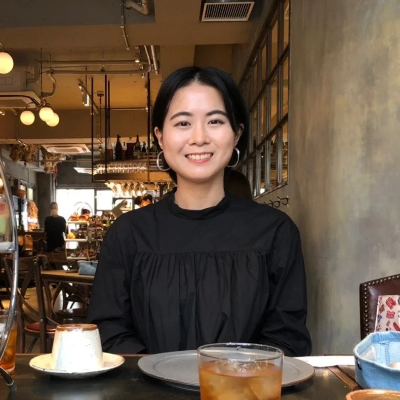 A woman with short black hair smiling at a restaurant table with a cup, a glass of iced tea, and a plate in front of her. The restaurant has a warm ambiance with hanging lights and a bar area in the background.