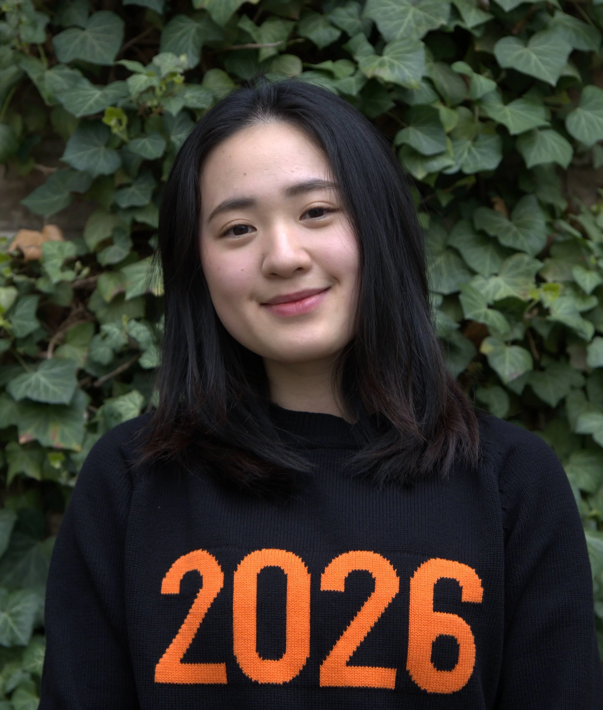 Portrait of a young woman with long dark hair, smiling, standing in front of green ivy leaves. She is wearing a black sweater with large orange numbers '2020' on the front.