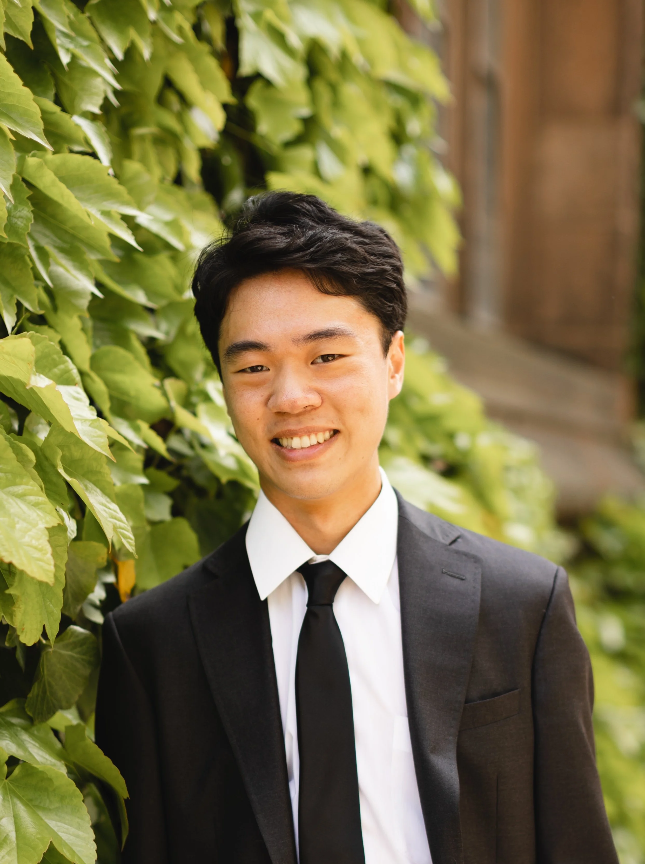 A young man in a black suit, white shirt, and black tie smiling outdoors near green foliage.