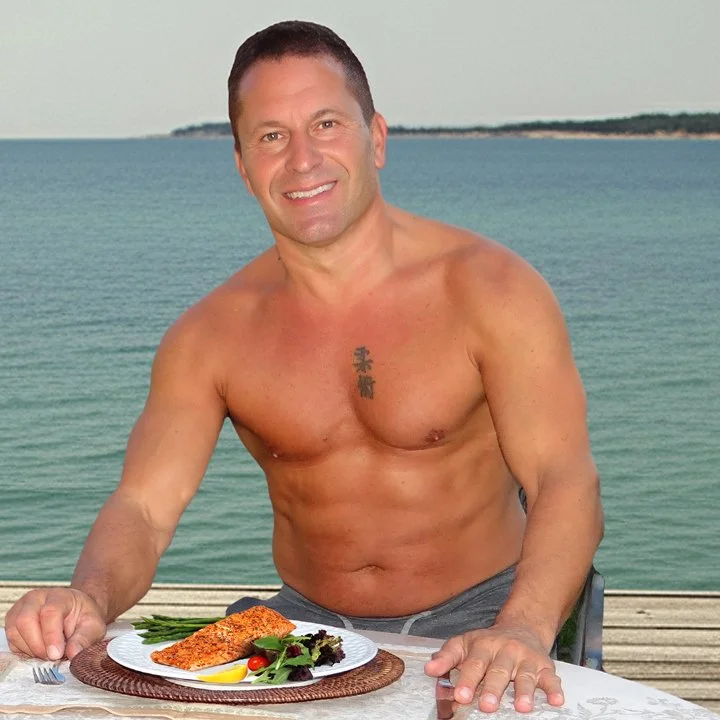 A shirtless man with short dark hair and tattoos on his chest, sitting at an outdoor table by the water, smiling, with a plate of food including fish, vegetables, and lemon in front of him.