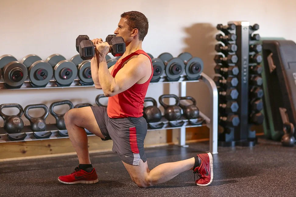 Man in a red tank top and shorts performs a lunge exercise with dumbbells in a gym.