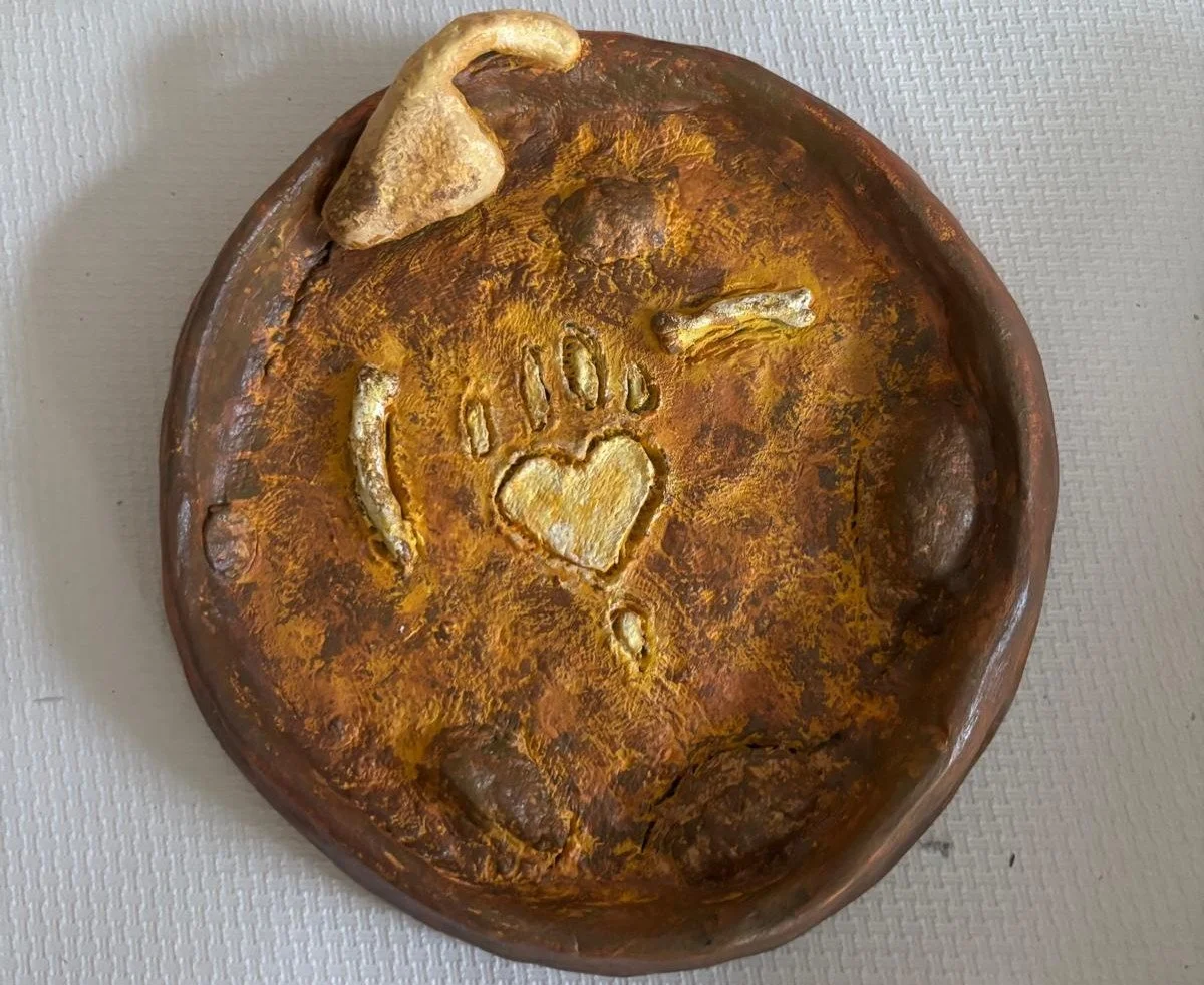 Manufatto in argilla decorato nei toni del marrone e del giallo, con una zampa d'orso stilizzata al centro, realizzato da uno dei partecipanti al laboratorio Manifattura Arcobaleno.