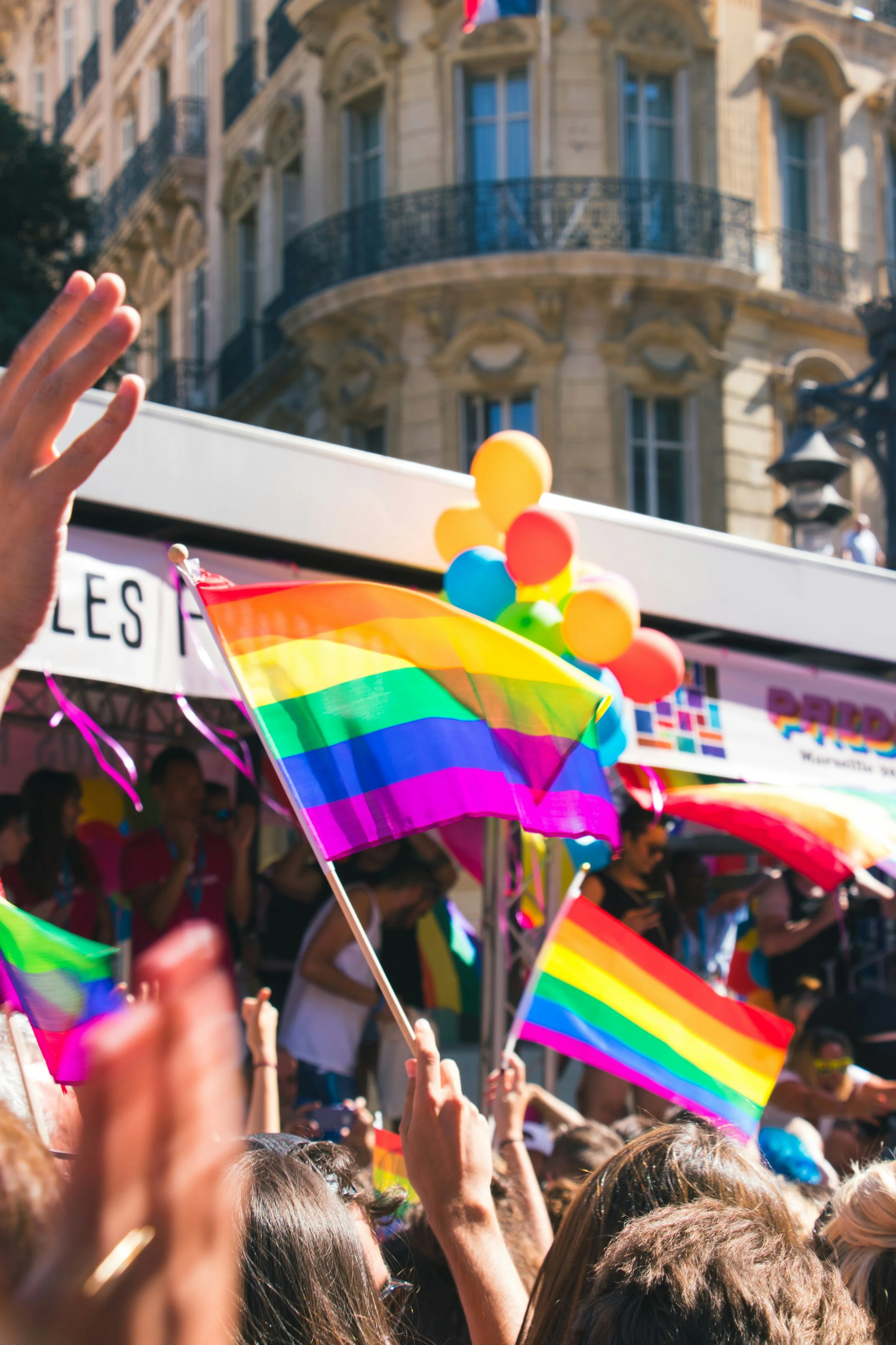 Persone al Pride con bandiere arcobaleno e palloncini colorati in strada di fronte a un edificio storico
