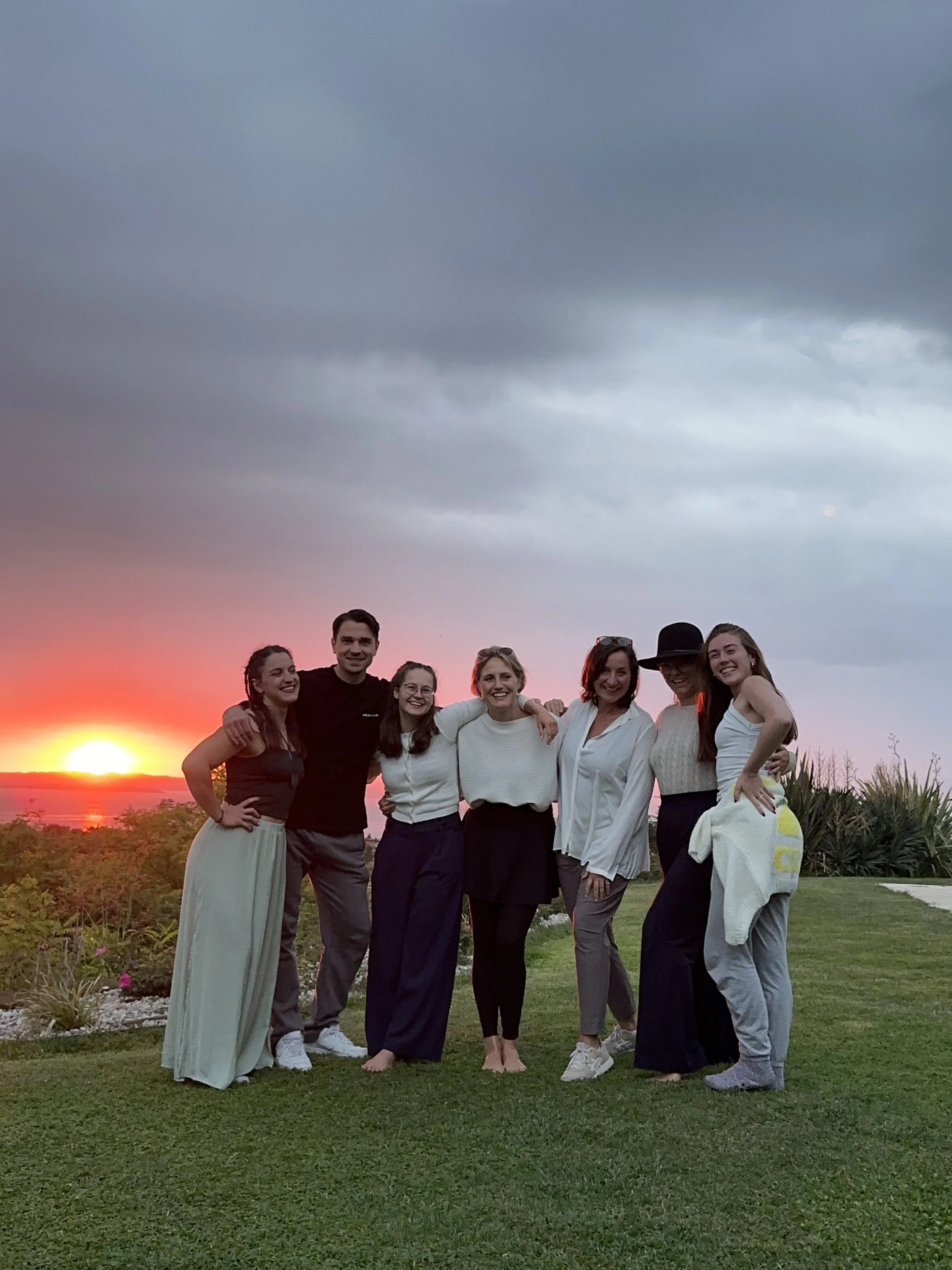 Eine Gruppe von acht jungen Menschen steht im Freien bei Sonnenuntergang, umarmt und lächelnd, auf grünem Rasen mit Büschen und Bäumen im Hintergrund.