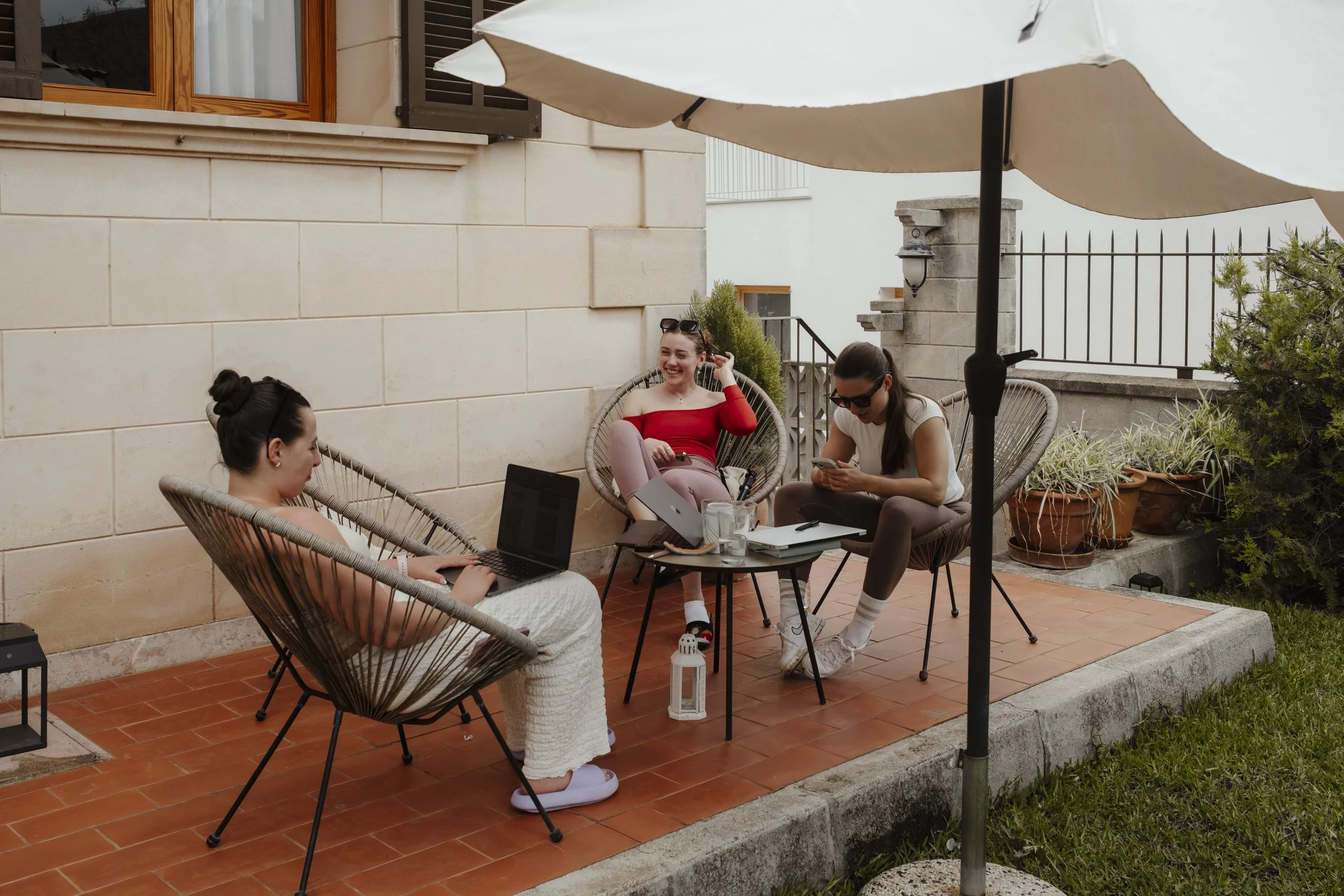Vier Frauen sitzen auf einem Terrassensofa unter einem Sonnenschirm, arbeiten auf Laptops und unterhalten sich, umgeben von Pflanzen.