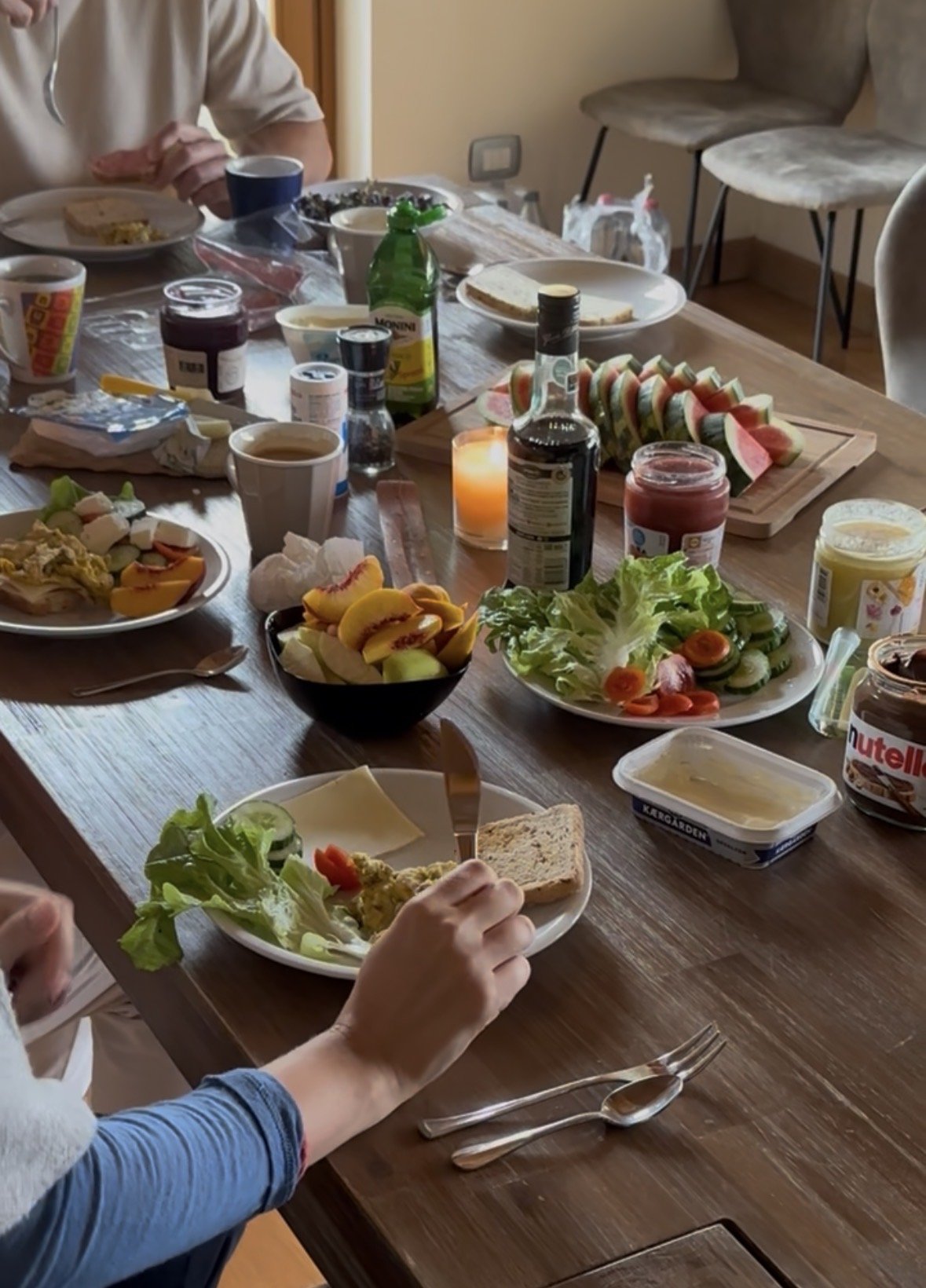 Ein Frühstückstisch mit verschiedenen Speisen, einschließlich Salat, Wurst, Obst, Marmelade, Nutella, Joghurt, Brot, Kaffee und Saft. Es sind Personen am Tisch, die essen oder servieren.