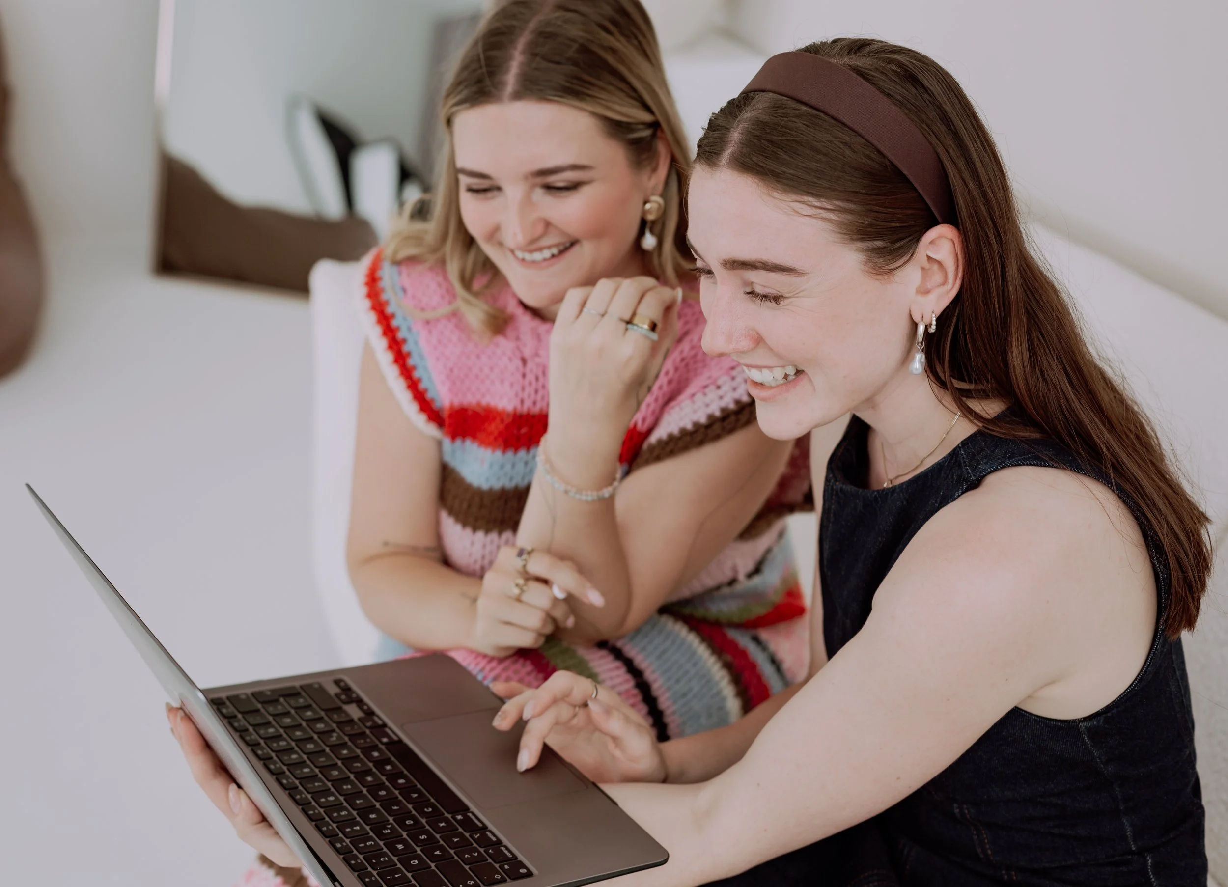 Zwei lachende Frauen schauen gemeinsam auf einen Laptop Bildschirm, während sie in einem modernen Raum sitzen.