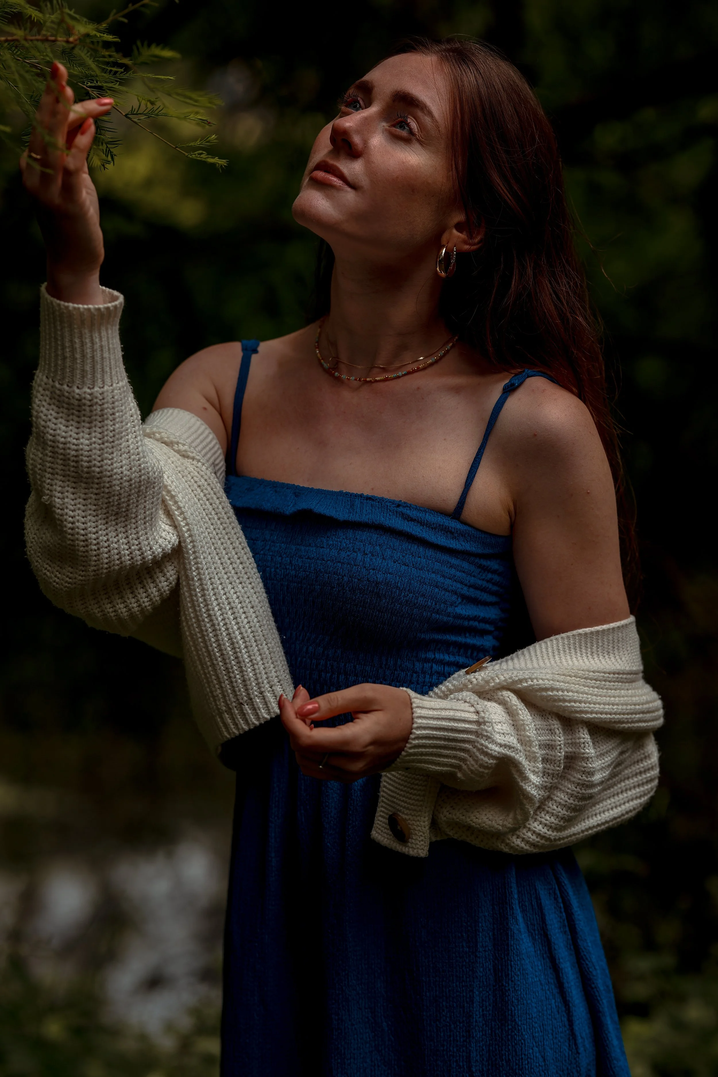 Eine Frau mit langen braunen Haaren trägt ein blaues Kleid und einen weißen, kuscheligen Cardigan, während sie eine Astgabel berührt, umgeben von einem Wald im Hintergrund.