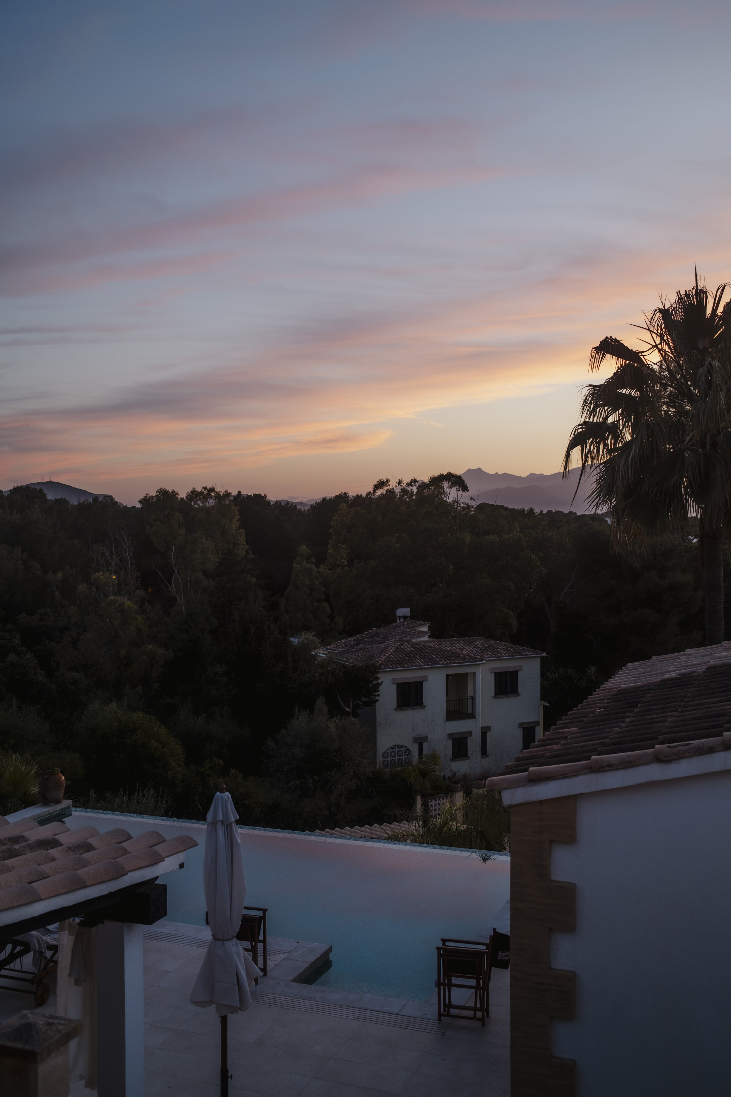 Blick auf den Sonnenuntergang über einem Wohngebiet mit Swimmingpool, umgeben von Bäumen und Bergen im Hintergrund.