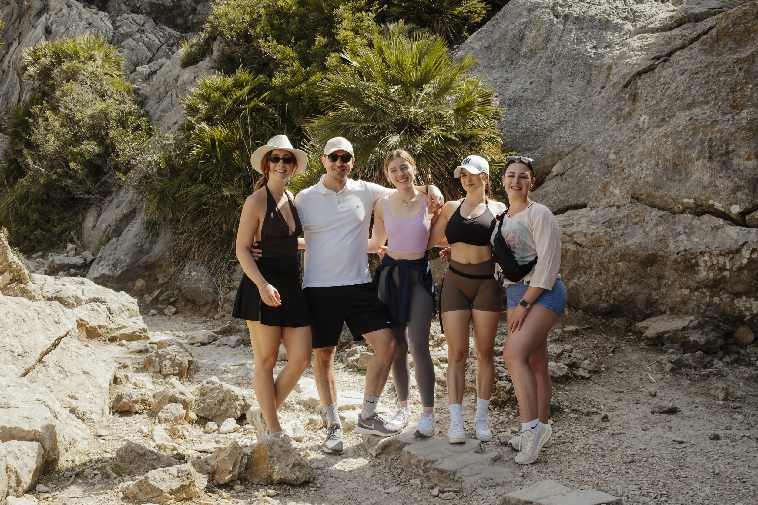 Fünf Menschen, die auf einem Felsen in einer trockenen, bergigen Landschaft stehen, lächeln und posieren für die Kamera. Es sind drei Frauen und zwei Männer, alle tragen sportliche Kleidung und Sonnenbrillen, einige mit Hüten.