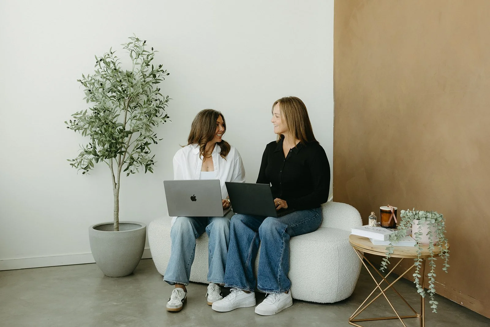 Two women sitting on a white couch, smiling at each other, with laptops on their laps, in a modern room with a tall potted plant and a side table with books, a plant, and decorative items.