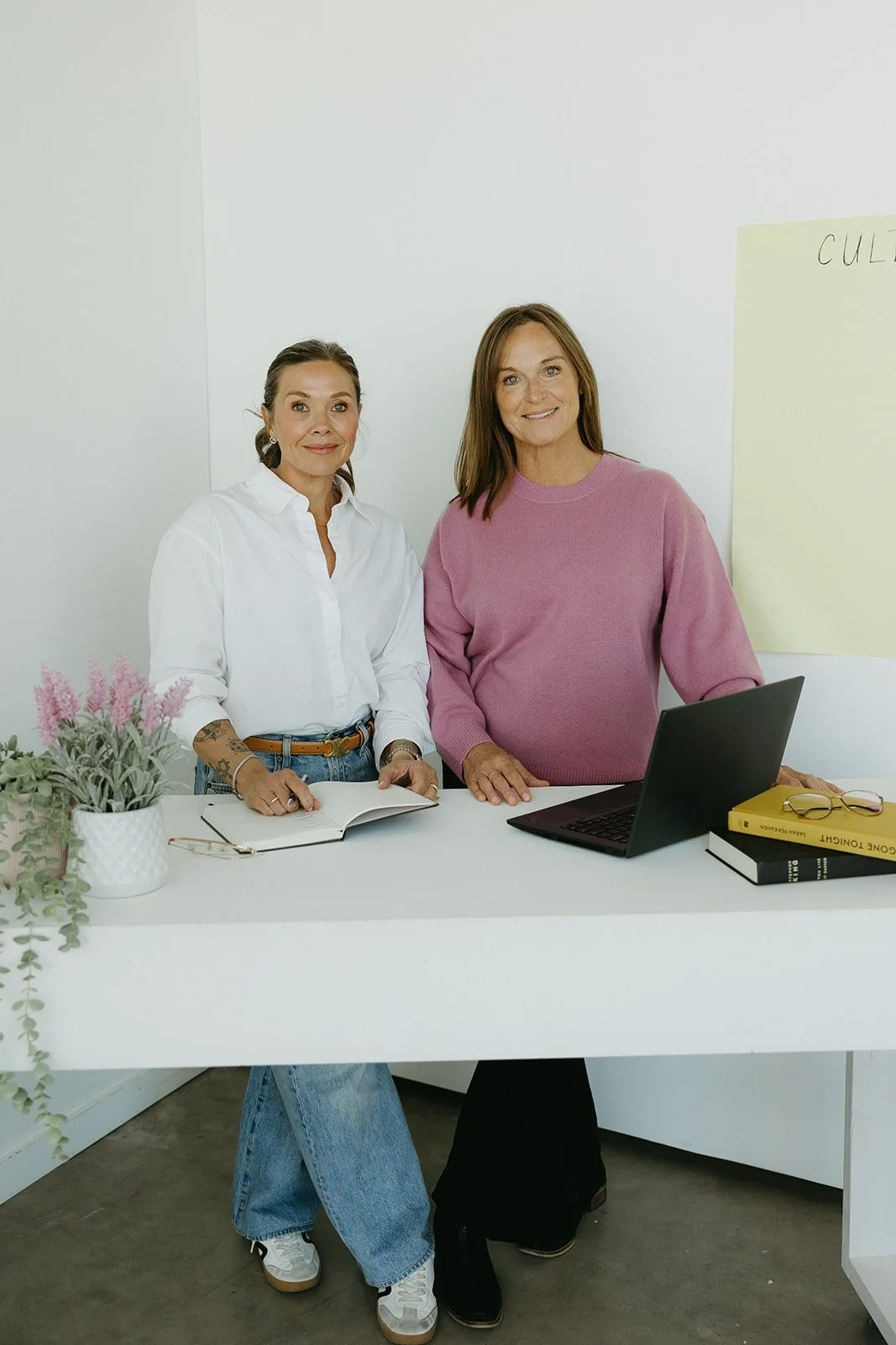 Two women standing behind a white desk, one wearing a white shirt and the other a pink sweater, in an office with a laptop, books, and a plant on the desk.