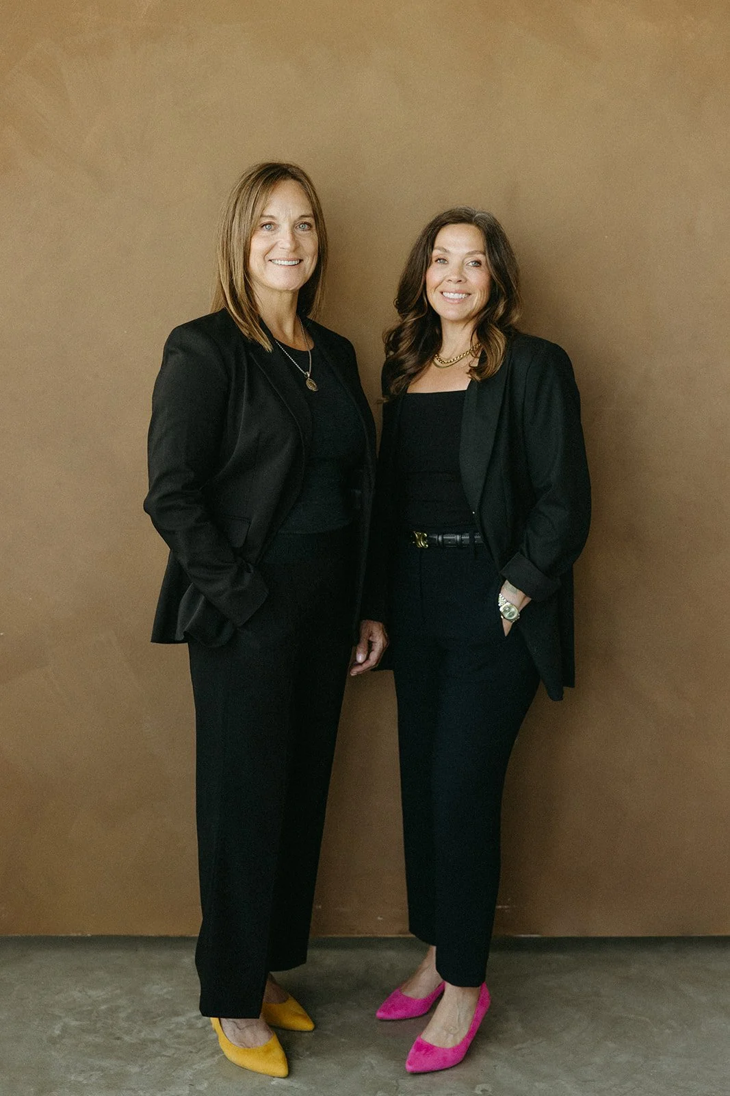 Two women standing side by side, dressed in black business attire, against a brown backdrop. One is wearing yellow heels, the other pink heels.