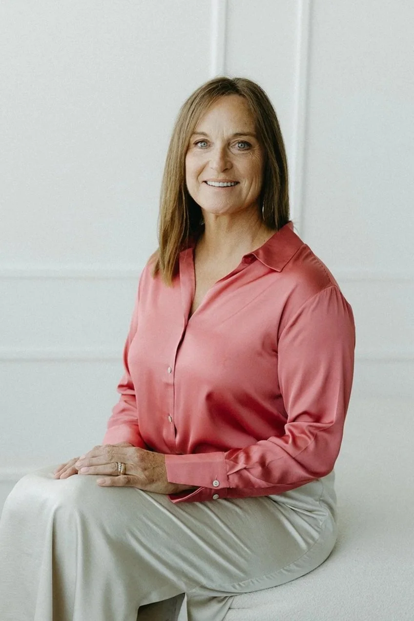 A woman with shoulder-length brown hair, blue eyes, and a warm smile, sitting on a white surface against a plain white background. She is wearing a pink satin button-up shirt and cream-colored pants.