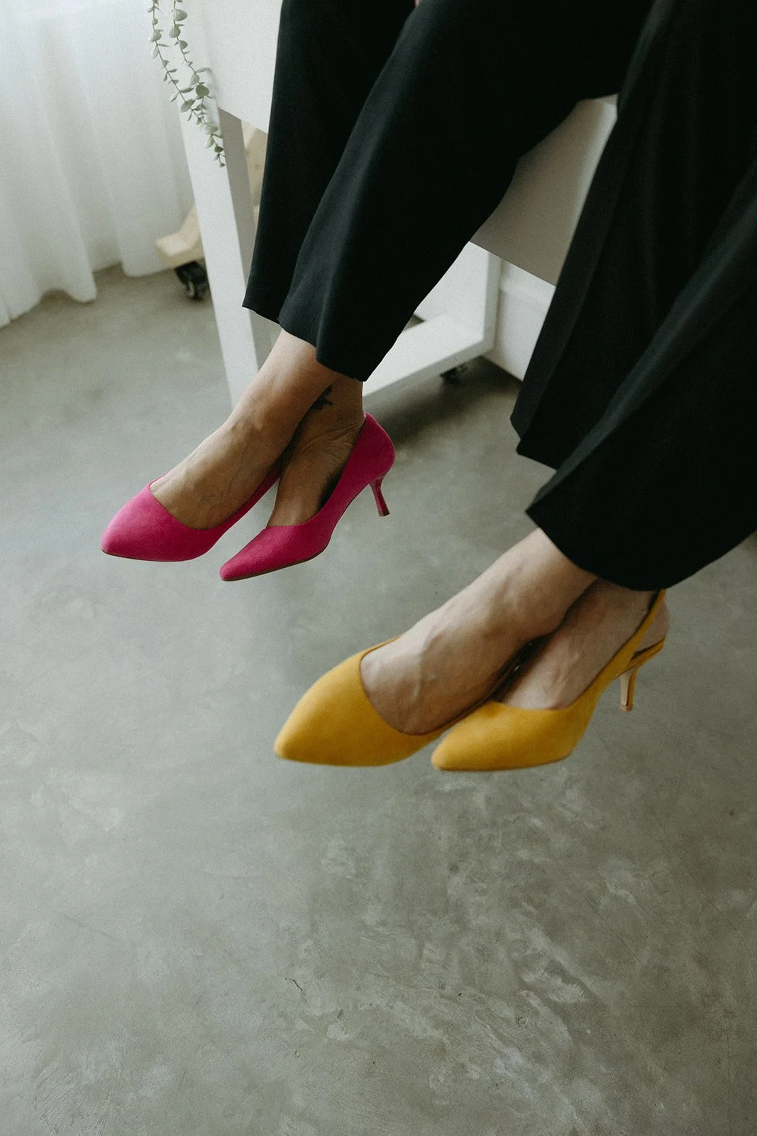 Person wearing black pants sitting on a white chair, wearing pink and yellow high-heeled shoes, with legs and feet extended downward.