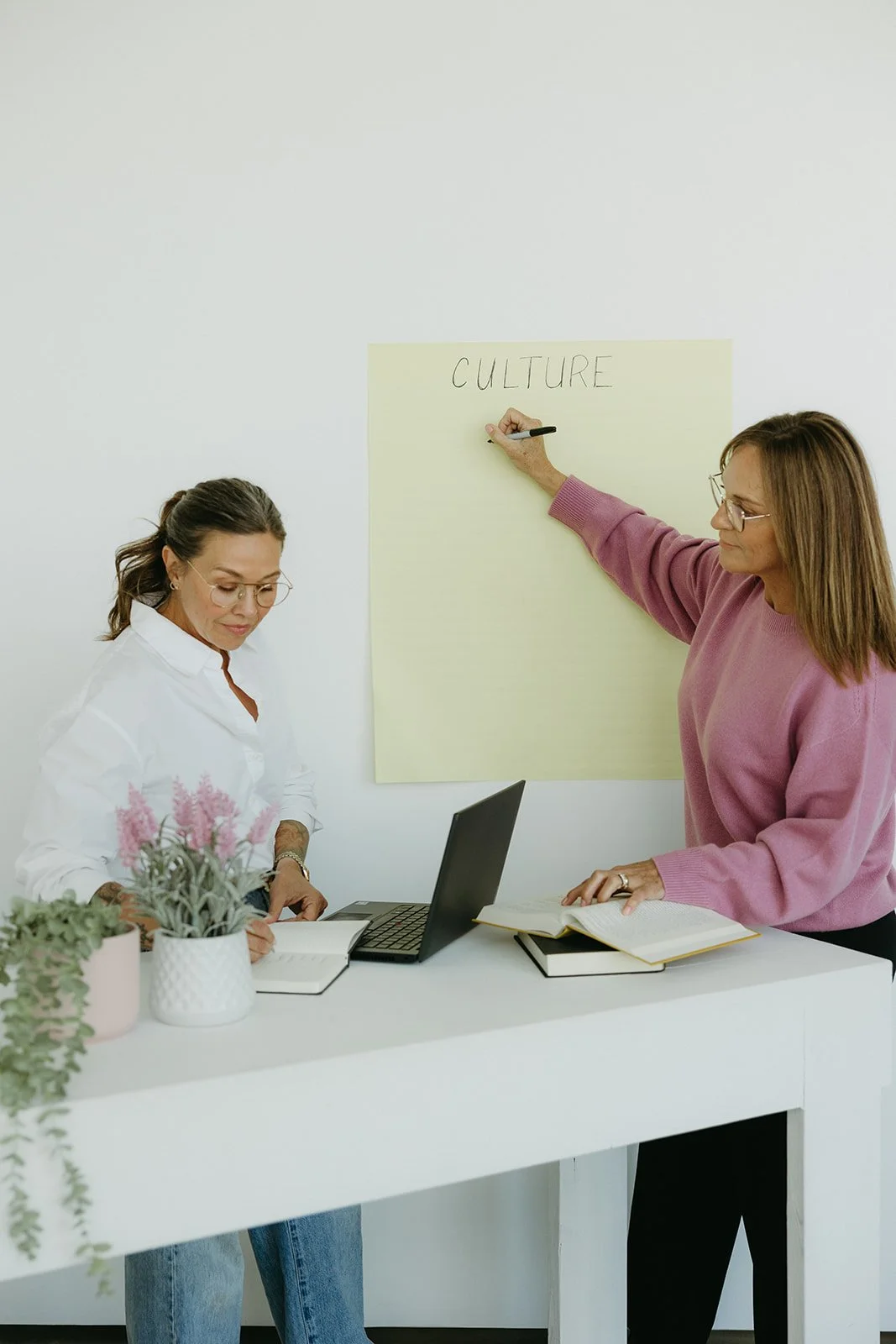 Two women in a meeting room, one is writing on a large yellow paper with 'CULTURE' written at the top, the other is standing with an open notebook, both are wearing glasses and casual clothes, there are notebooks, a laptop, and potted plants on the table.