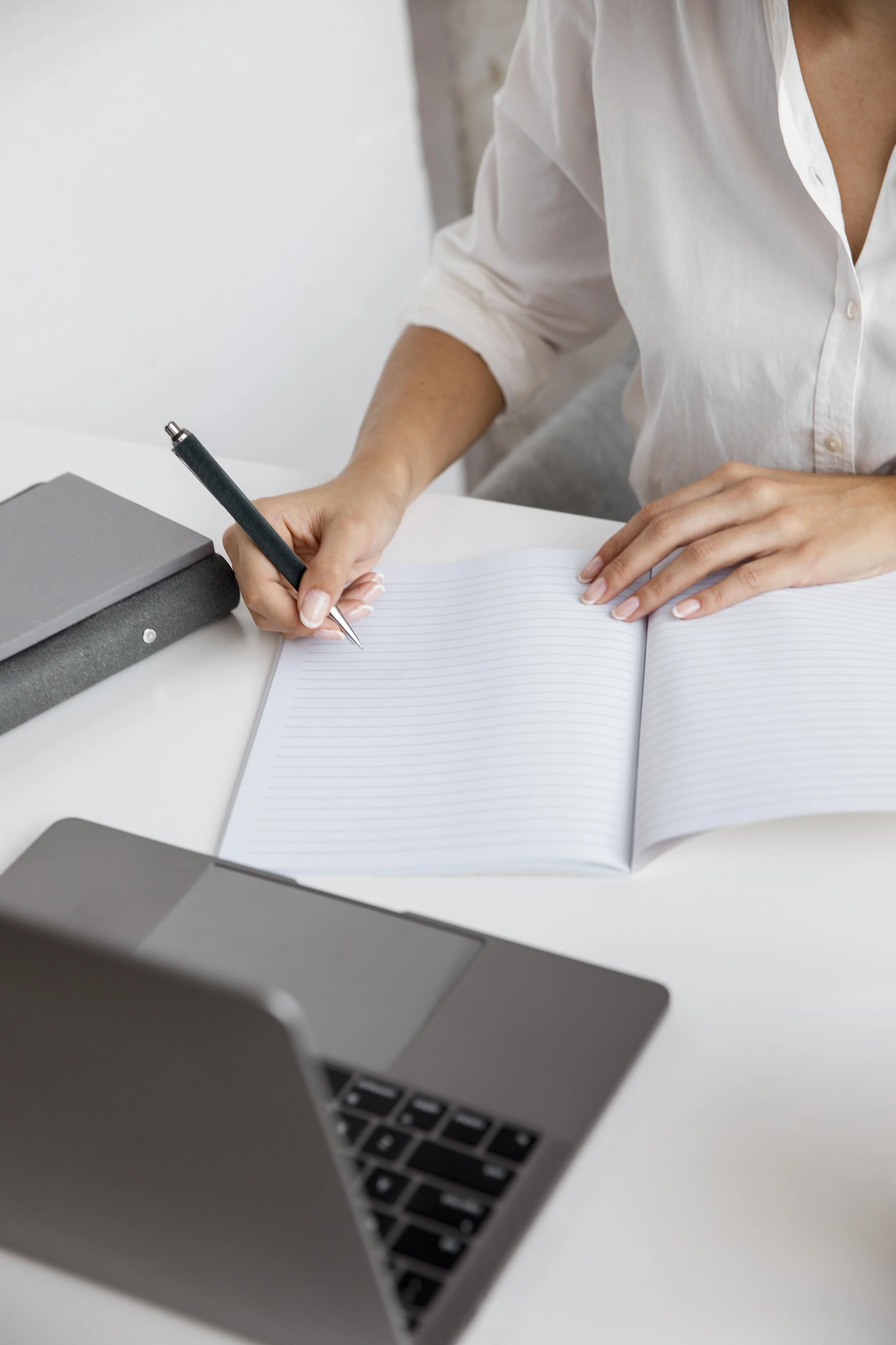Person writing in a lined notebook with a black pen, sitting at a white desk with a closed laptop and gray notebooks nearby.