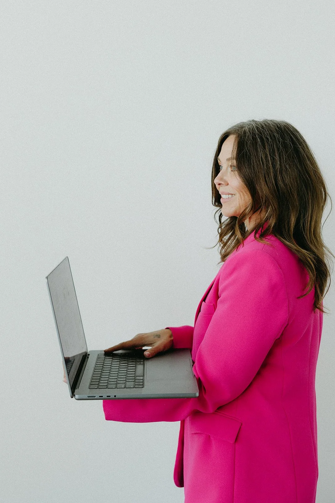 Woman in bright pink blazer holding open laptop and smiling, facing right, against a plain white background.