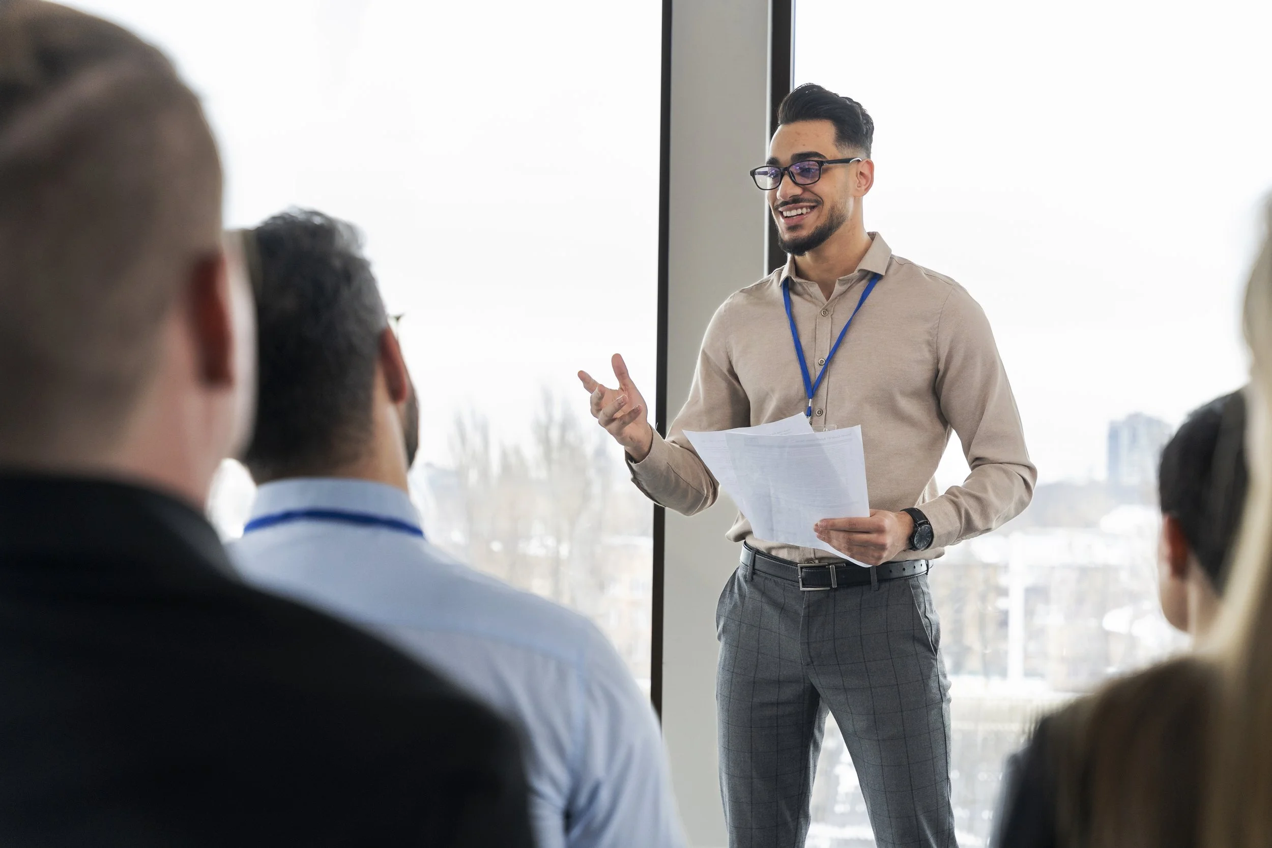 A man with glasses and a beige shirt giving a presentation to a group of people in a conference room with large windows.