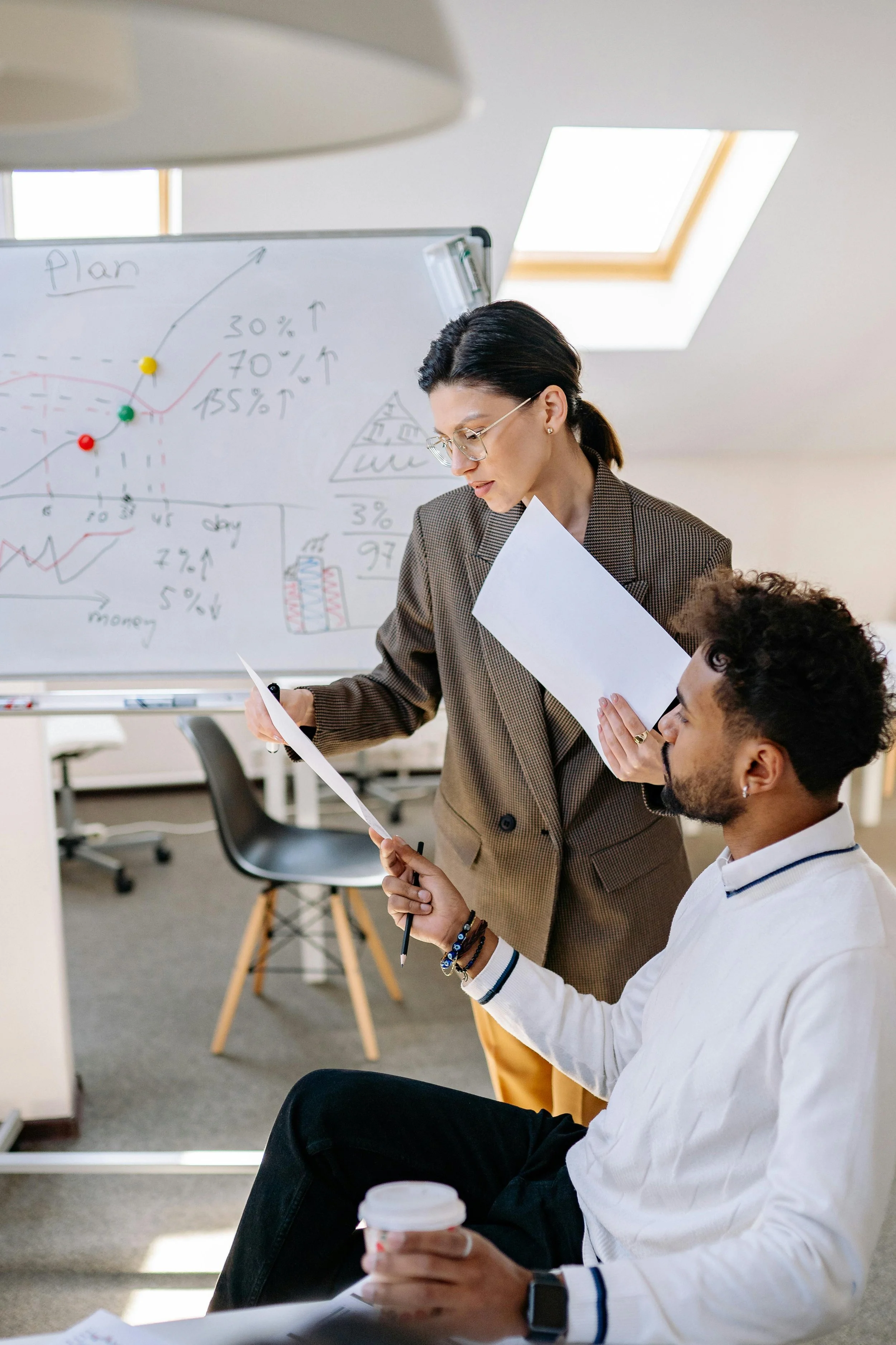 Two professionals in an office discussing data, with a whiteboard filled with charts and graphs in the background.