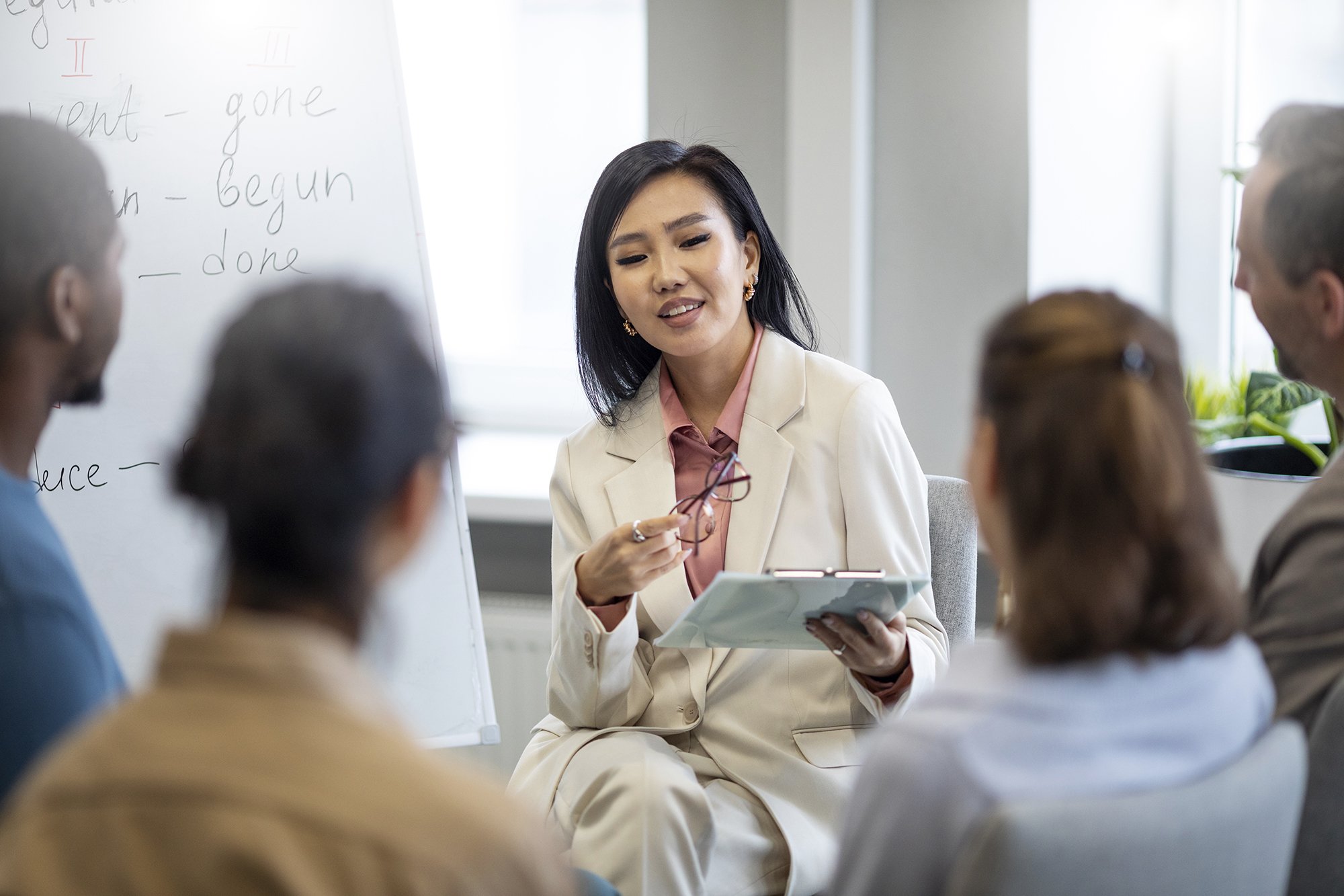 A woman in a white suit teaching a class or holding a discussion with a group of people in a classroom or office setting, with a whiteboard in the background.