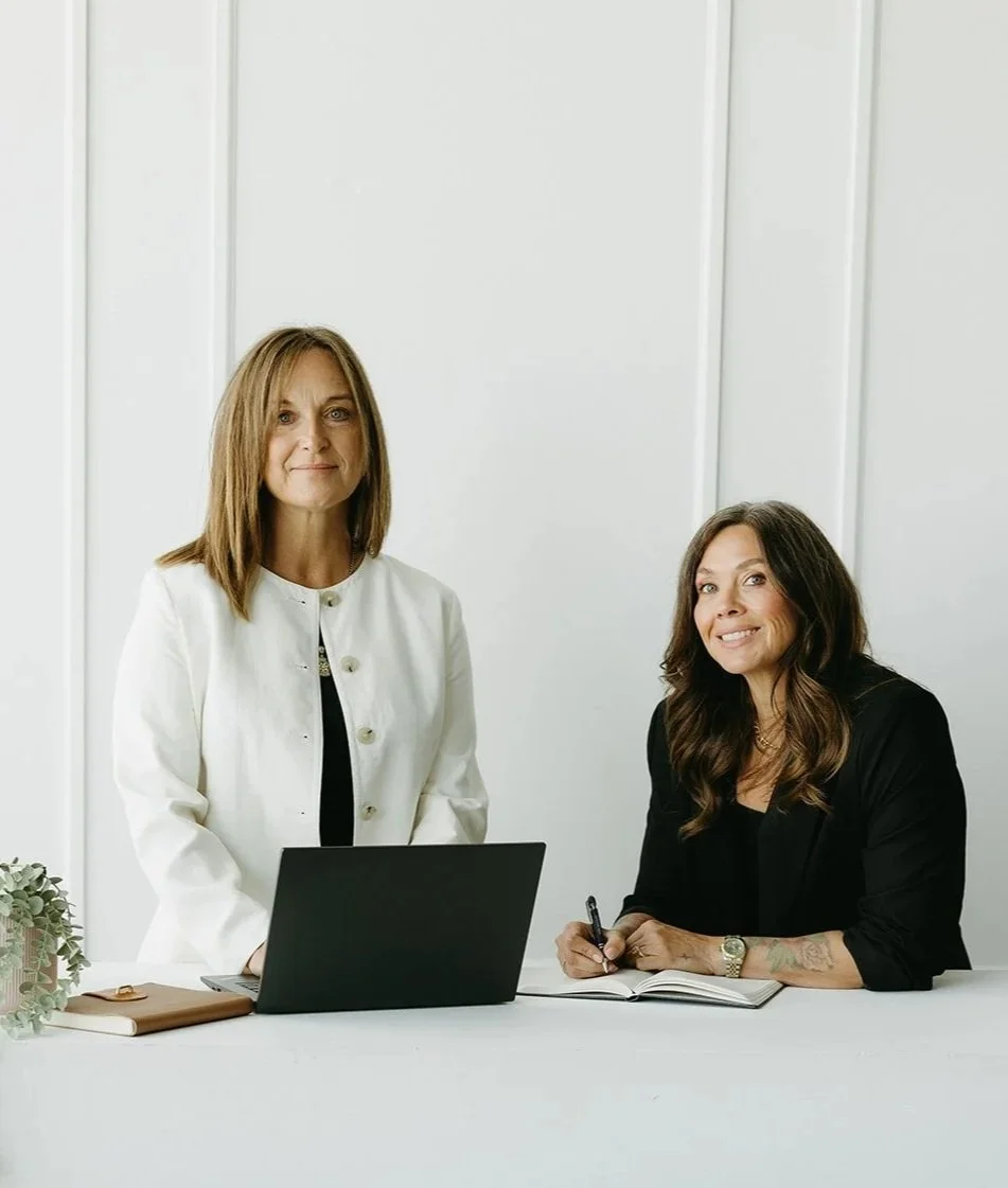Two women in professional attire, one standing and one sitting at a white table with a laptop, notebook, and plant in a bright, modern office.