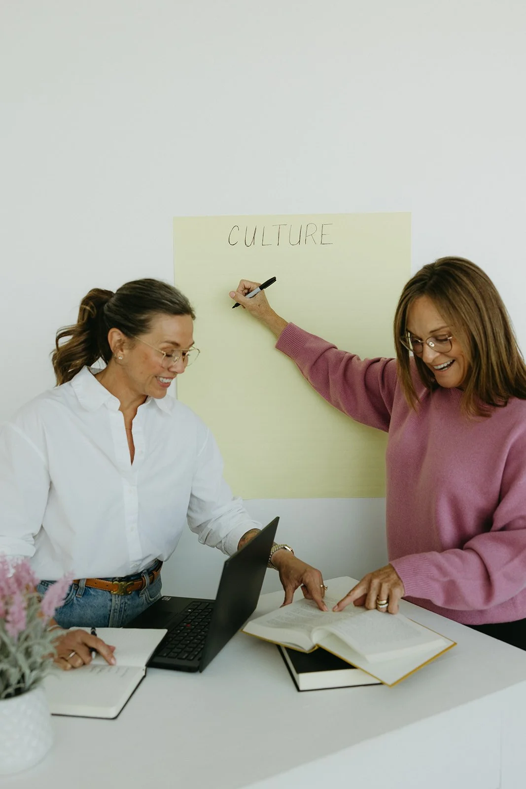 Two women working together at a desk, smiling, with a laptop, notebooks, and a book open. One woman points at an open book while the other looks on. A large yellow paper on the wall behind them has 'CULTURE' written on it.