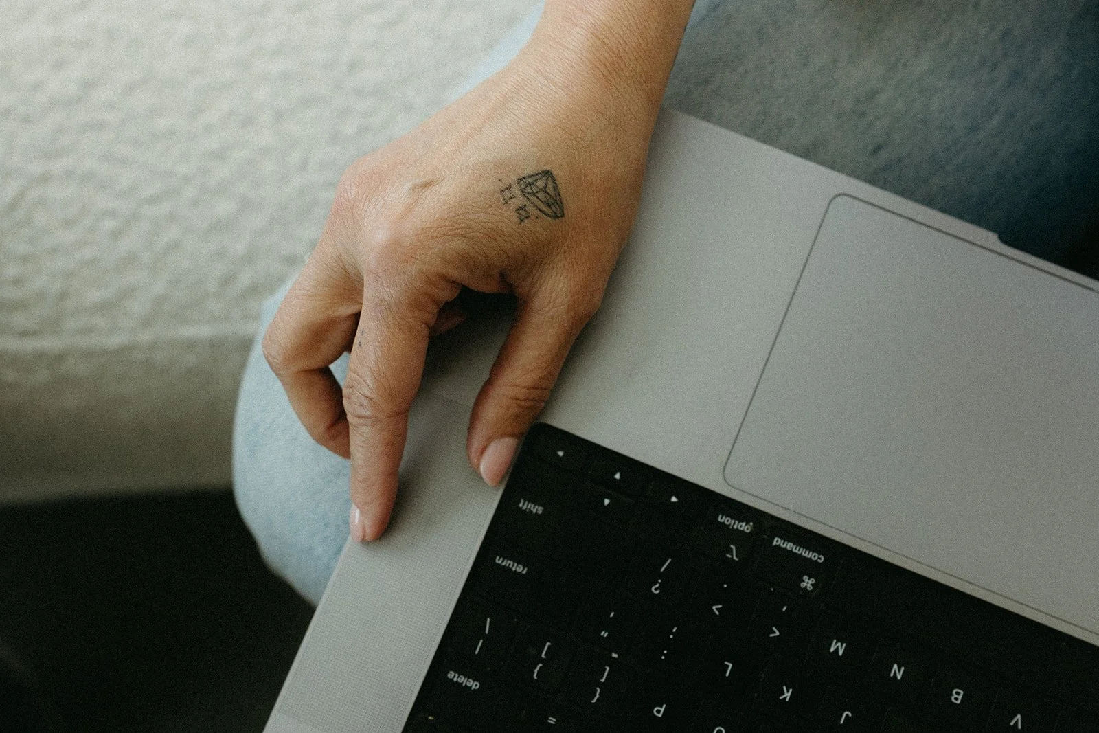 A person with a small diamond tattoo on the hand, holding the corner of a silver laptop with a black keyboard, seated on a gray surface.