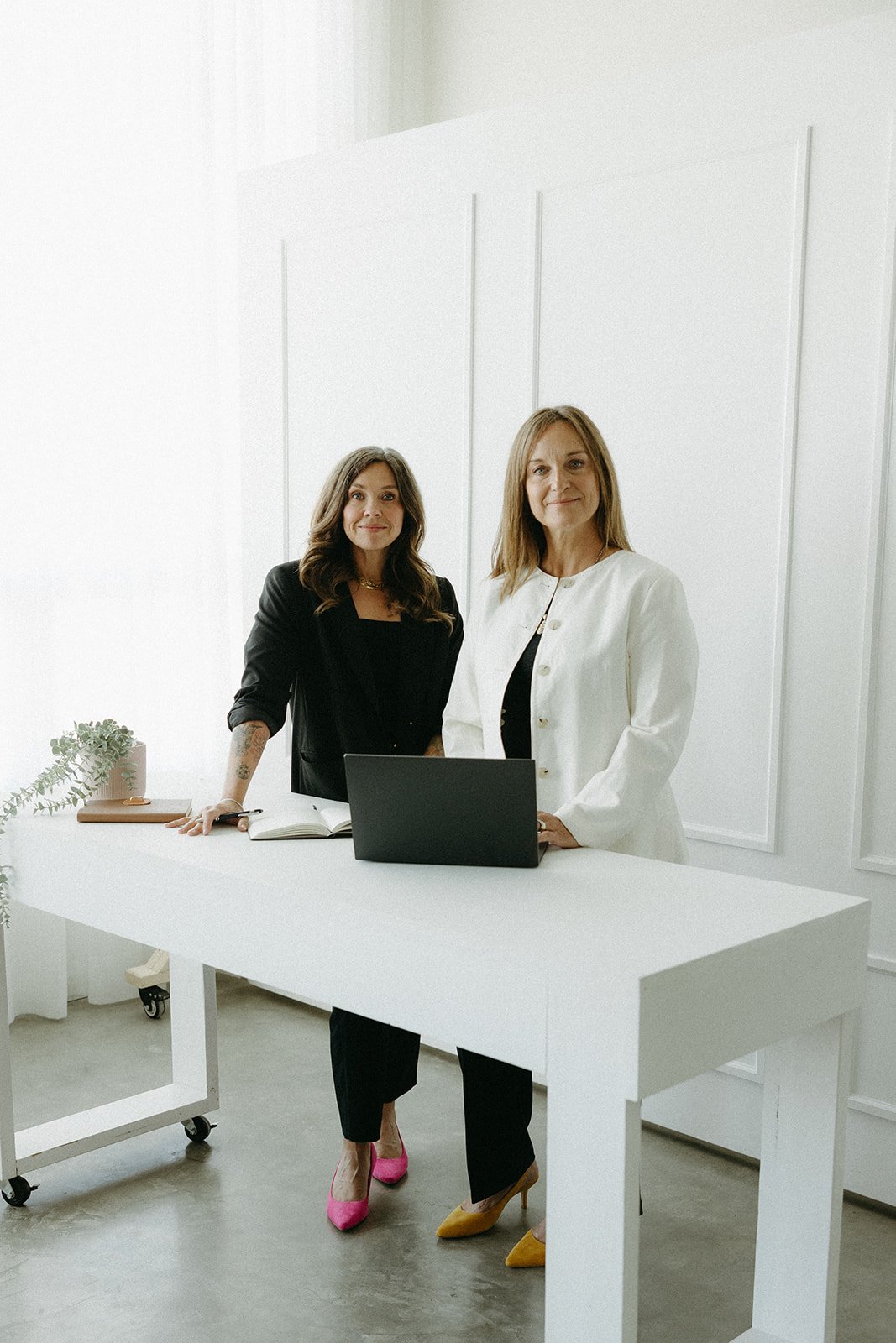 Two women standing behind a white table in an office space with a laptop, open notebook, and plant on the table, smiling at the camera.
