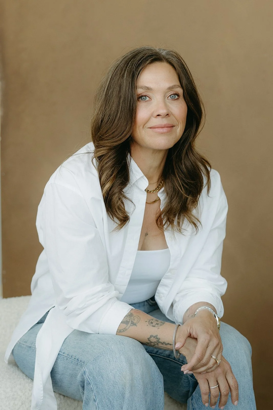 A woman with long brown hair, wearing a white shirt and blue jeans, sitting with folded hands, looking at the camera, with a neutral brown background.