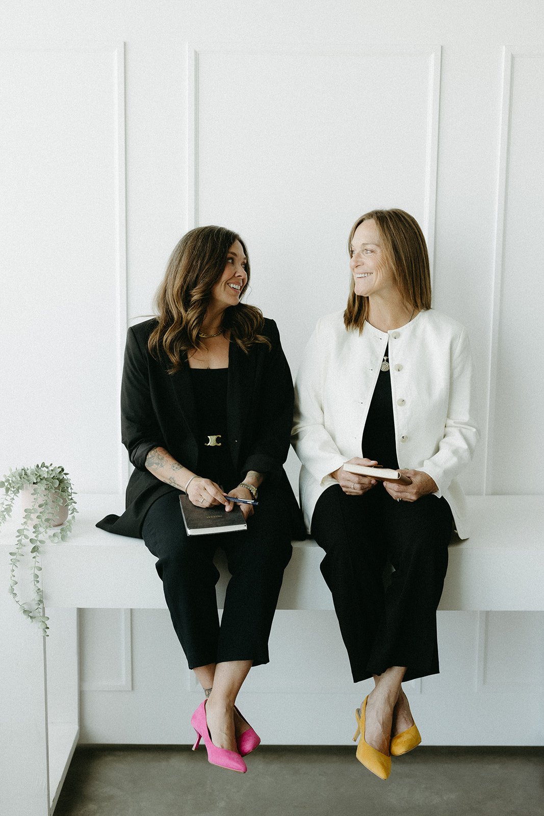 Two women sitting on a white bench, smiling and looking at each other, dressed in black and white, with bright pink and yellow high heels.