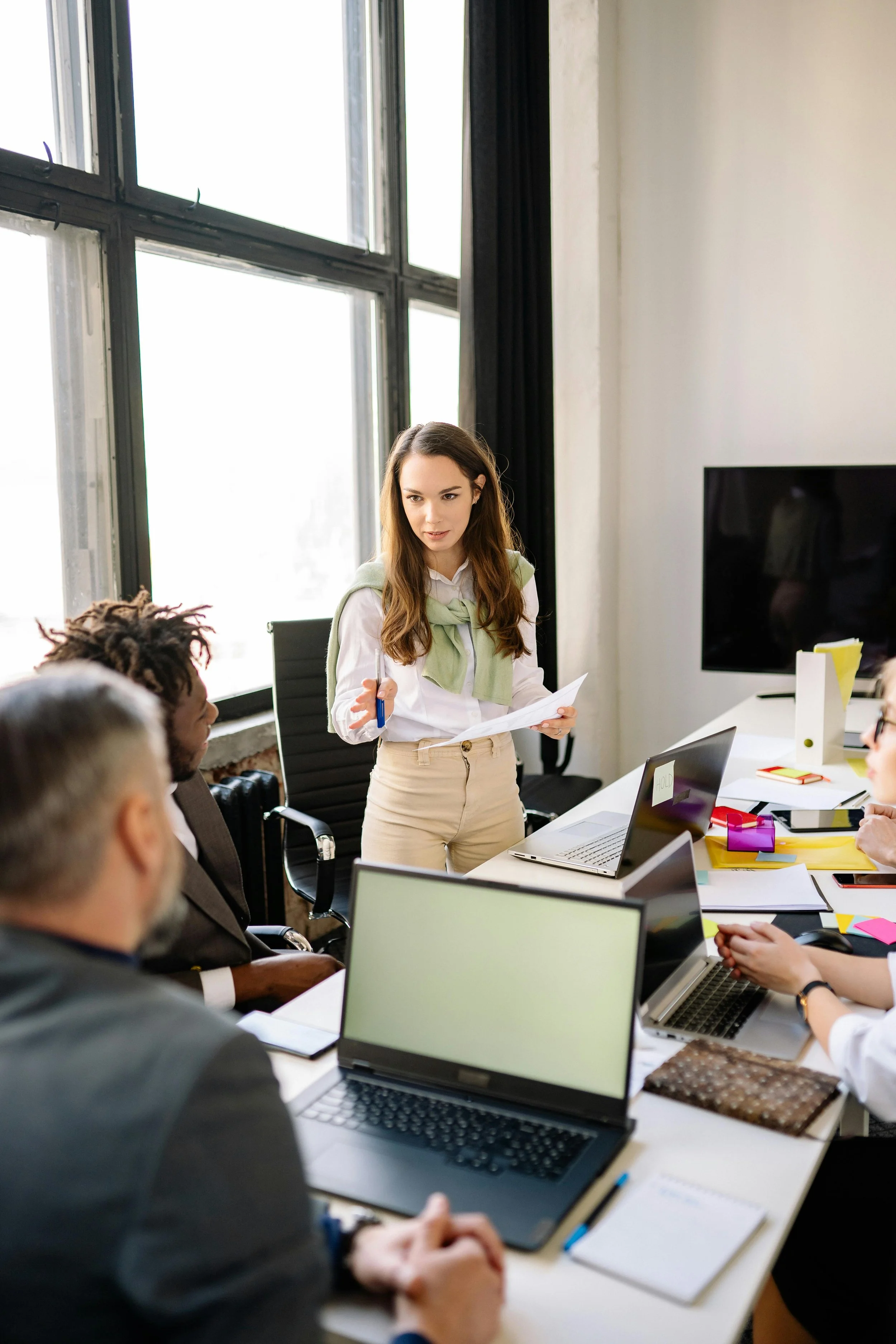 A woman presenting to a group of people in a meeting room with laptops and documents on the table, near large windows.