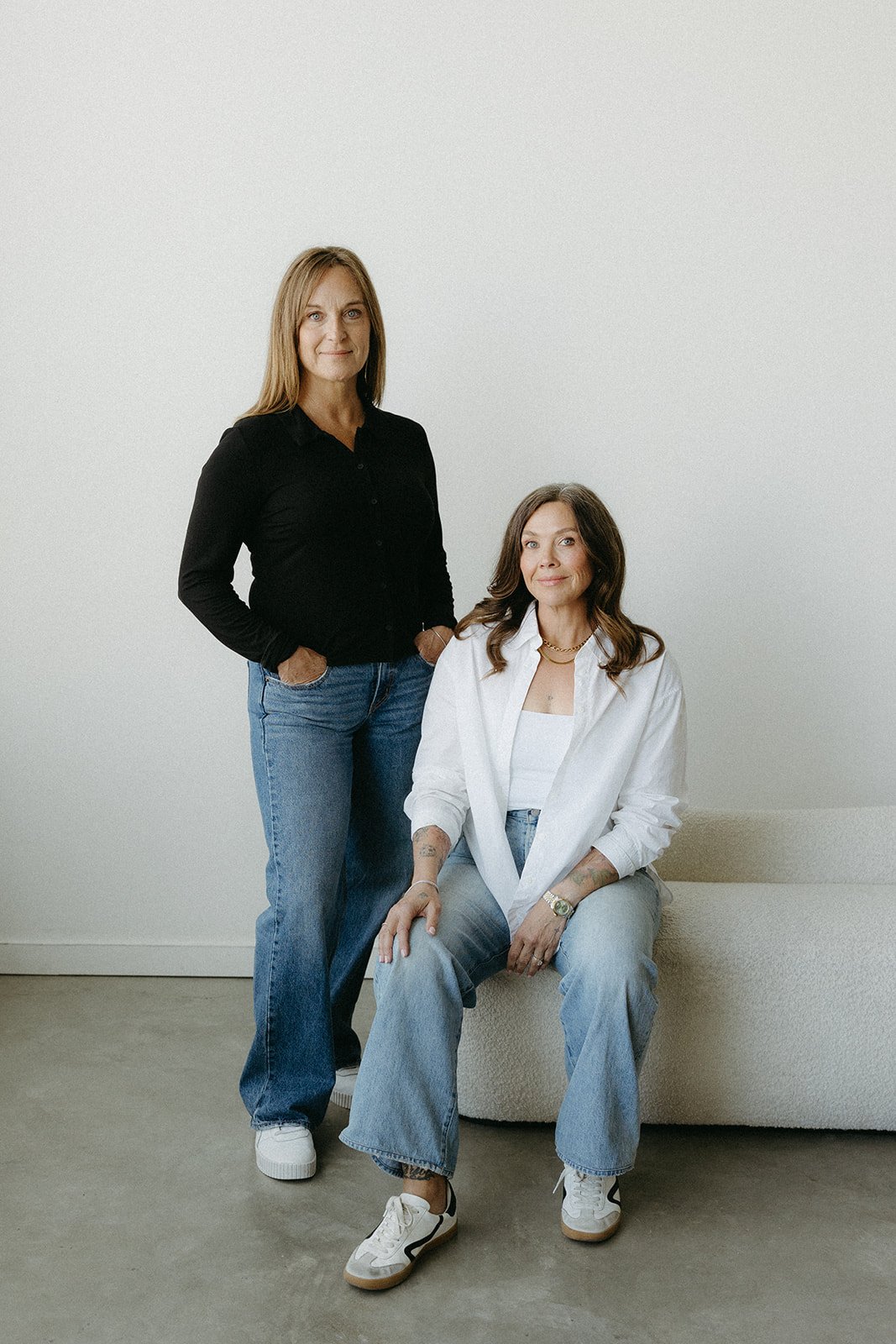 Two women with casual clothing, one sitting and one standing, in a minimalistic room with a white wall background.