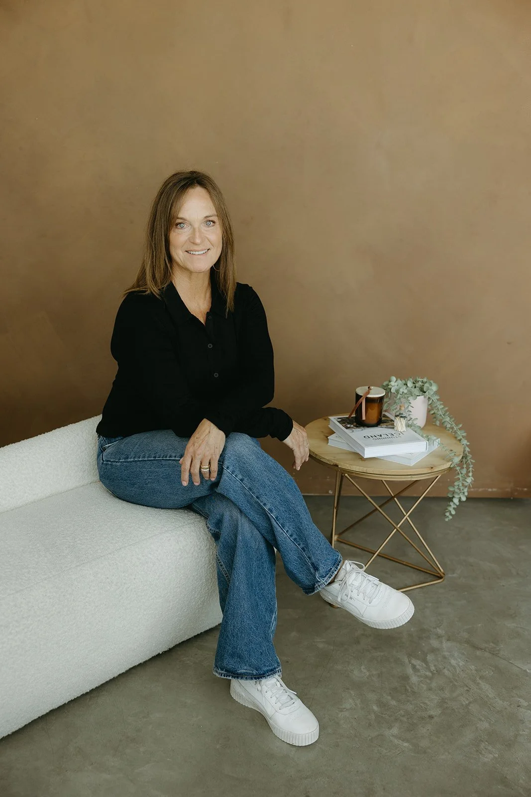 A woman sitting on a beige sofa with crossed legs, wearing a black shirt, blue jeans, and white sneakers, smiling at the camera. There is a round coffee table with magazines, a mug, and a white vase with green foliage beside her.