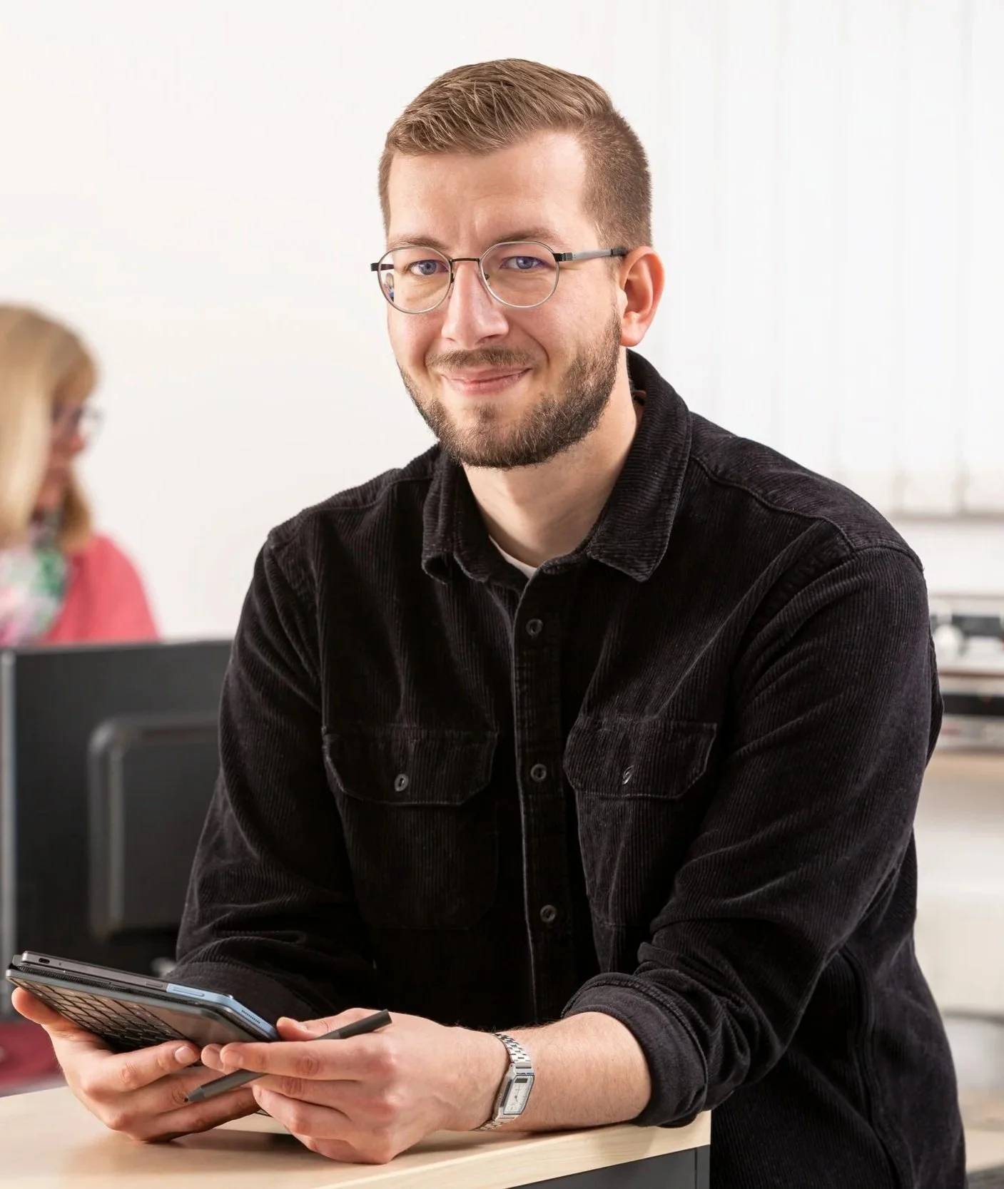 Junge Mann mit Brille und dunklem Hemd sitzt an einem Tisch, hält ein Tablet und lächelt in die Kamera. Im Hintergrund sind andere Personen im Büro zu sehen.