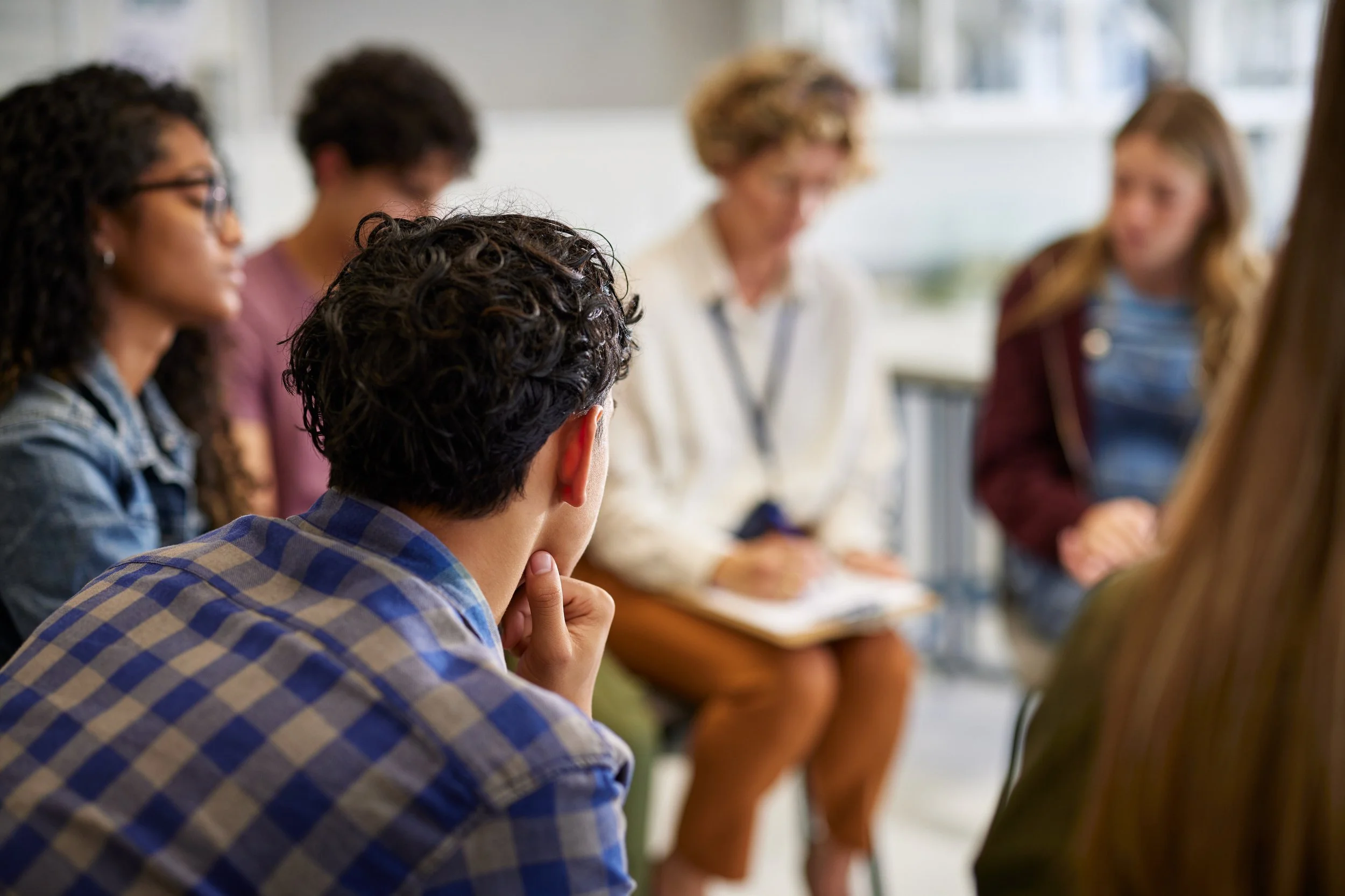 Group of diverse young adults participating in a support group in a bright room, with a woman with curly hair and glasses speaking to the group.
