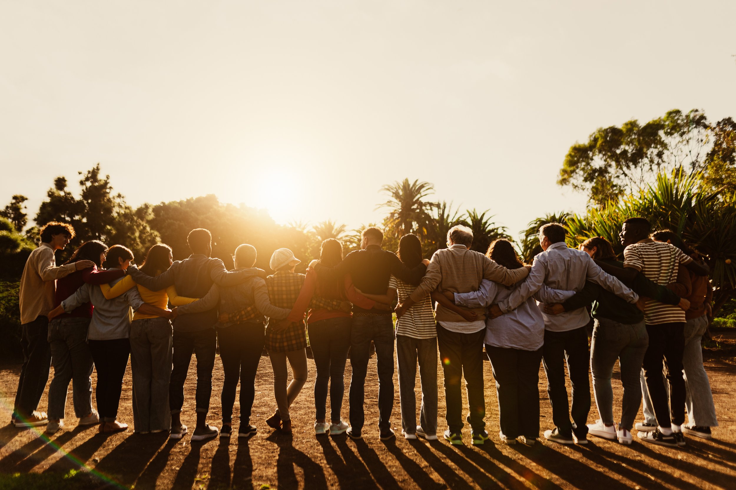 A diverse group of people standing in a circle with arms around each other's shoulders, outdoors at sunset, with trees and plants in the background.