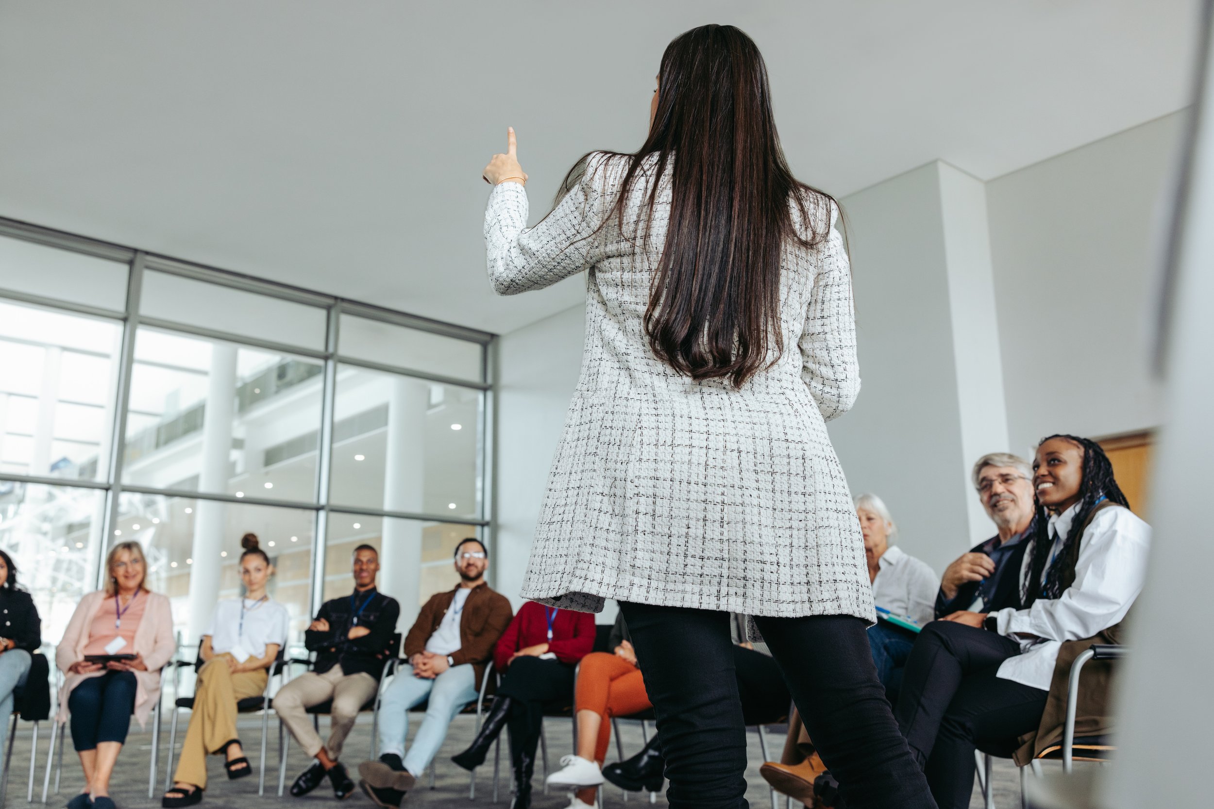 A woman in a patterned blazer speaks to an audience of diverse adults seated in a circular arrangement in a modern conference room, with large windows and natural light.