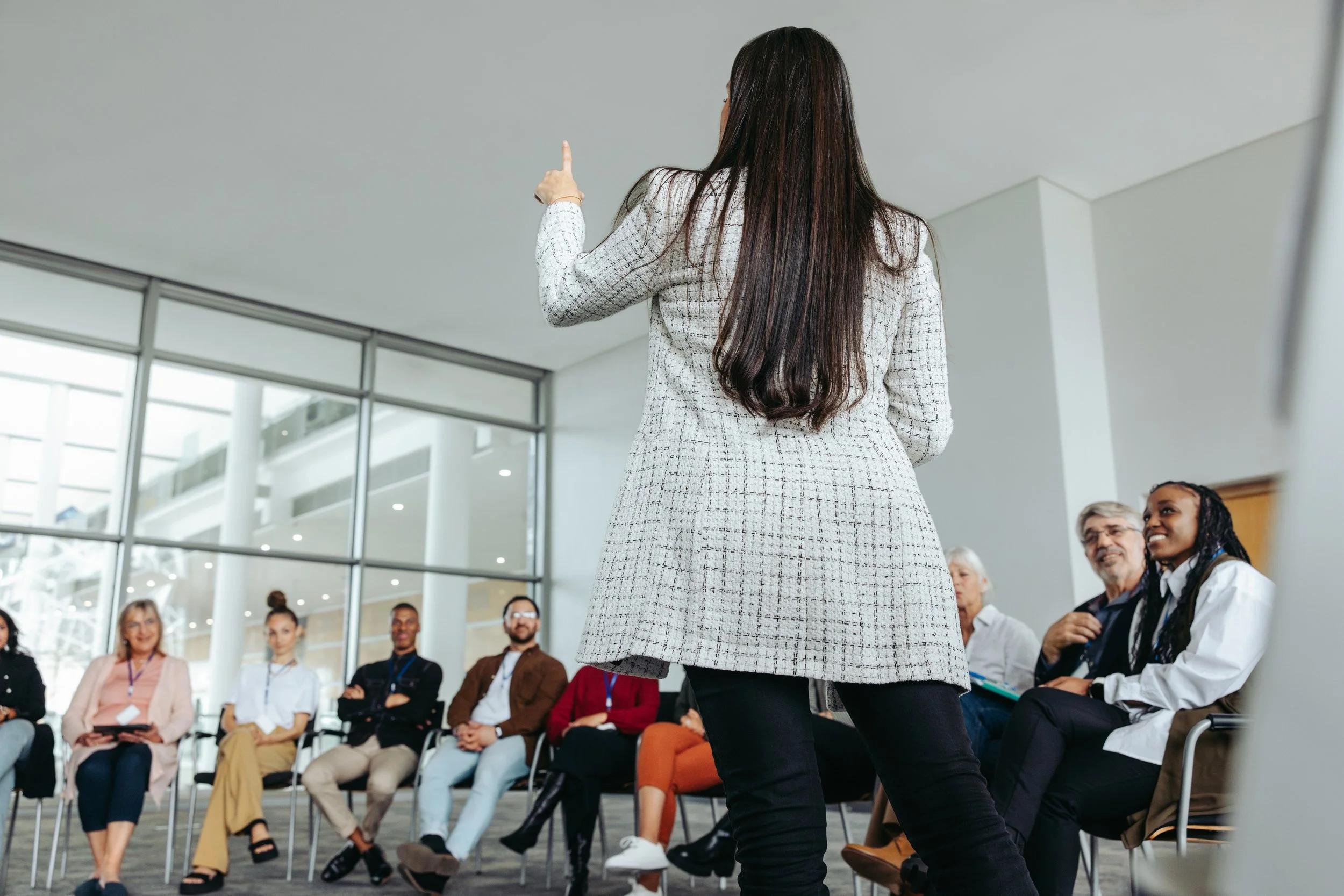 A woman in a patterned blazer speaks to an audience of diverse adults seated in a circular arrangement in a modern conference room, with large windows and natural light.