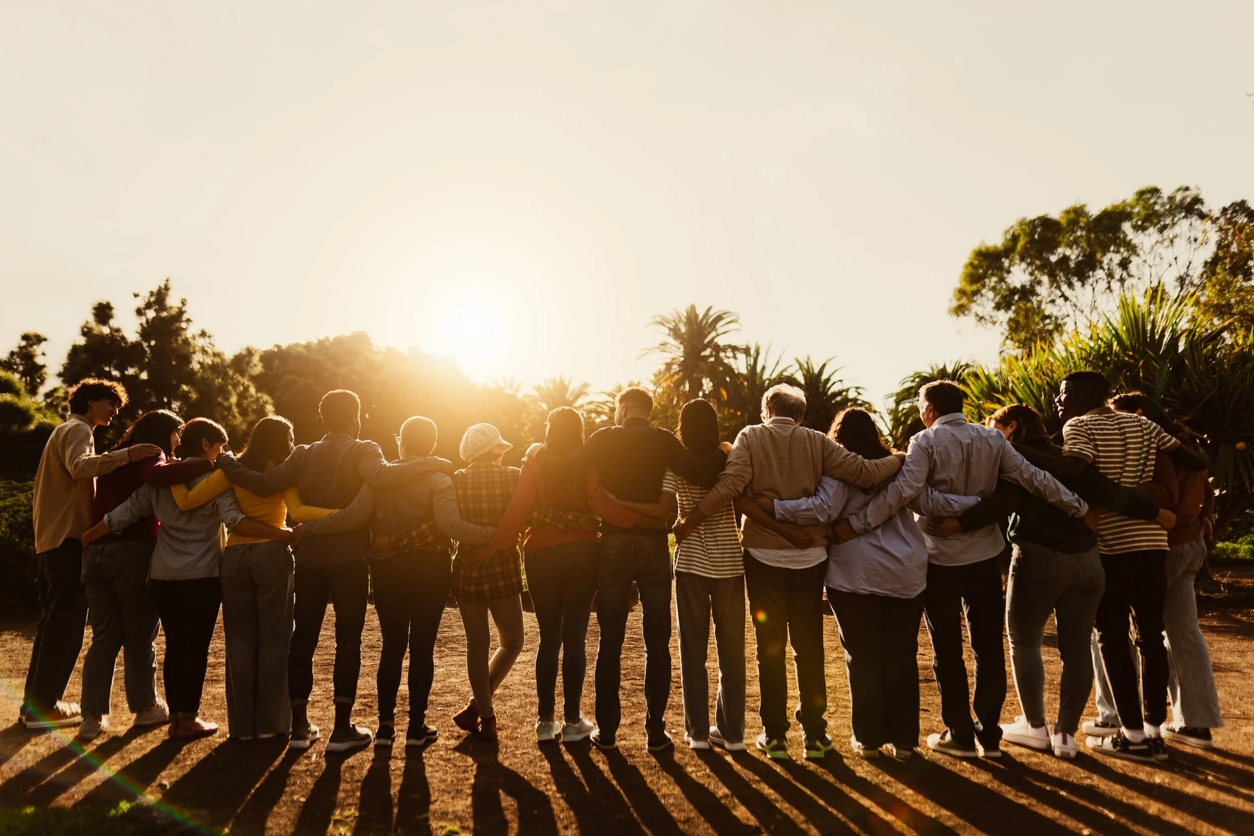 A diverse group of people standing in a circle with arms around each other's shoulders, outdoors at sunset, with trees and plants in the background.