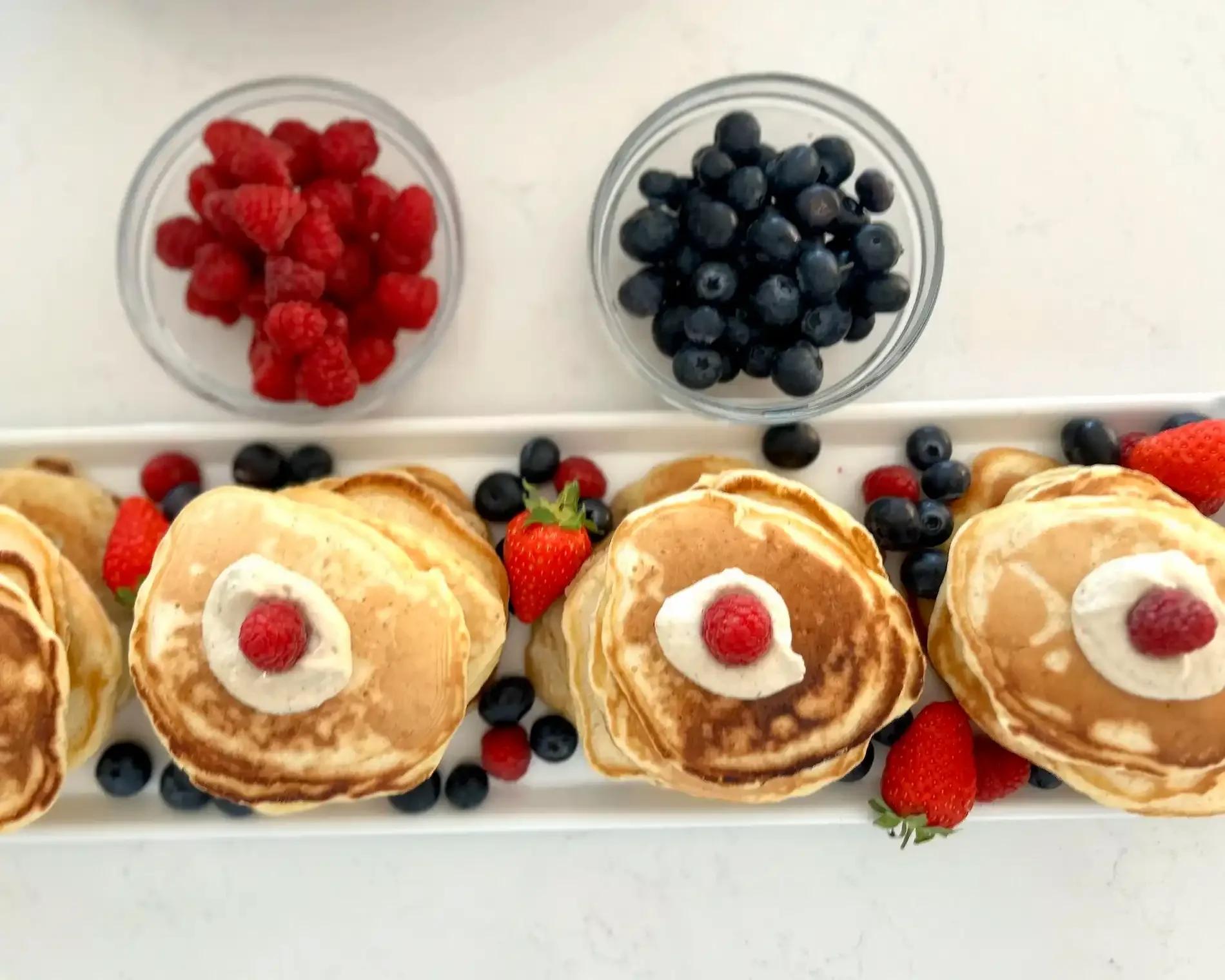 A plate of five pancakes topped with raspberry and served with strawberries and mixed berries, with additional berries in two small bowls.