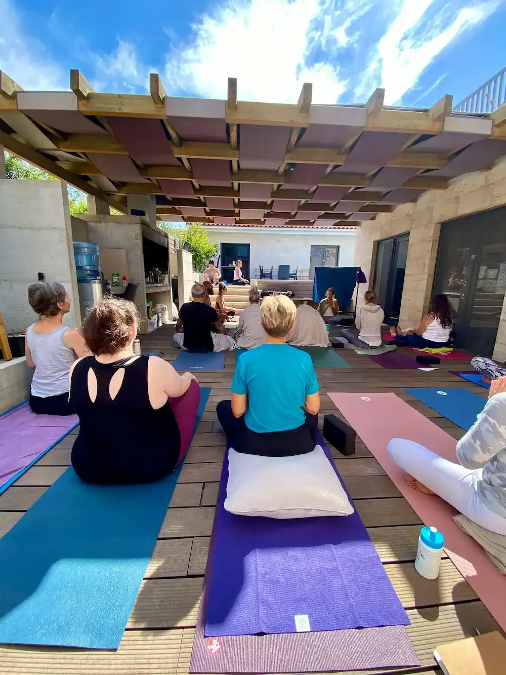 People participating in a yoga class on an outdoor deck, seated on yoga mats with their backs to the camera, facing a instructor at the front. The deck is covered by a wooden structure and surrounded by glass doors and concrete walls.