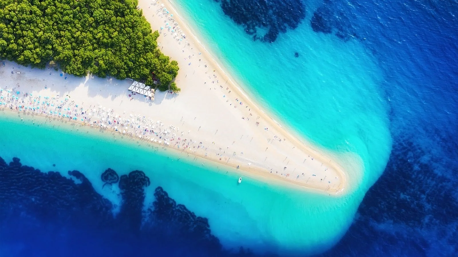 Aerial view of a sandy beach on an island with turquoise waters, surrounded by dark blue ocean, with crowded umbrellas and a dense green forest area.